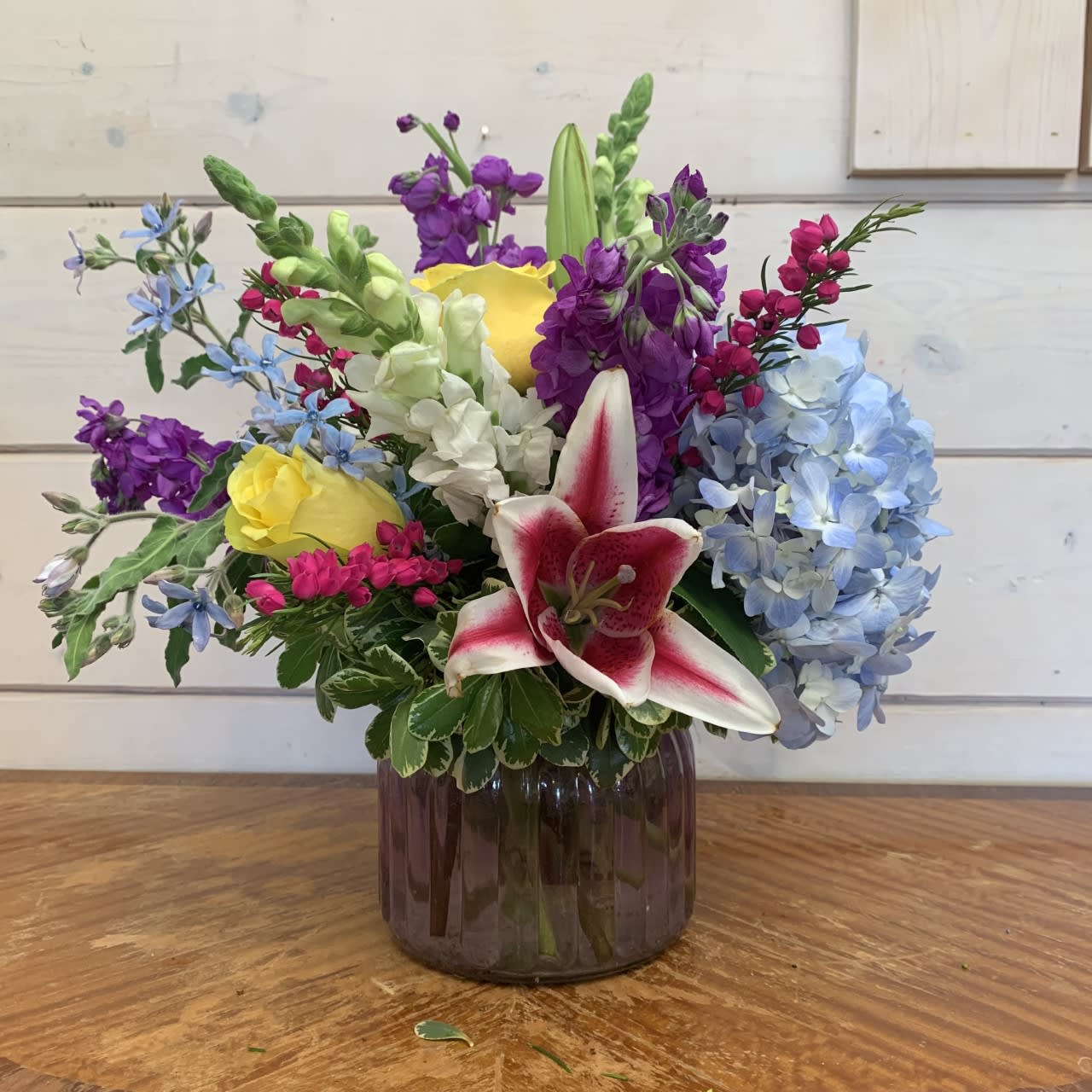Cheerful Greetings  - blue hydrangea, purple stock, yellow roses, white snapdragons, and stargazer lilies arranged in a extra large purple ginger jar with tweedia, beronia or other seasonal filler flowers. Approximately 14in tall and 12in wide.   