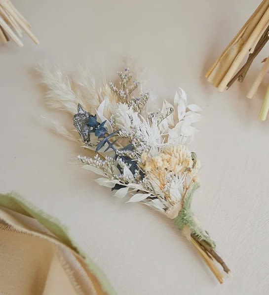 Boutonnière Fleurie Séchée Ivoire - An elegant dried boutonniere in ivory tones, enhanced with pampas grass, preserved eucalyptus, white nigella, limonium, baby’s breath, and white ruscus. Perfect for Prom, Homecoming, weddings or any formal event seeking a timeless touch. 