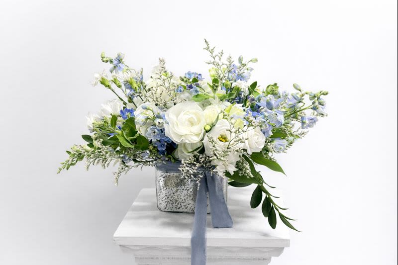 Blue Reverie - Blue Reverie – Blue &amp; White Floral Arrangement  A soft and elegant blue and white floral arrangement inspired by the “something blue” wedding tradition. Designed with premium white blooms, delicate blue florals, and fresh greenery, this arrangement symbolizes love, harmony, and new beginnings.  Handcrafted in a timeless glass vase and finished with a flowing ribbon, it’s ideal for weddings, bridal celebrations, engagements, baby celebrations, baby showers, anniversaries, or refined everyday gifting.  Florals may vary slightly based on seasonal availability while maintaining the overall color palette and design aesthetic.