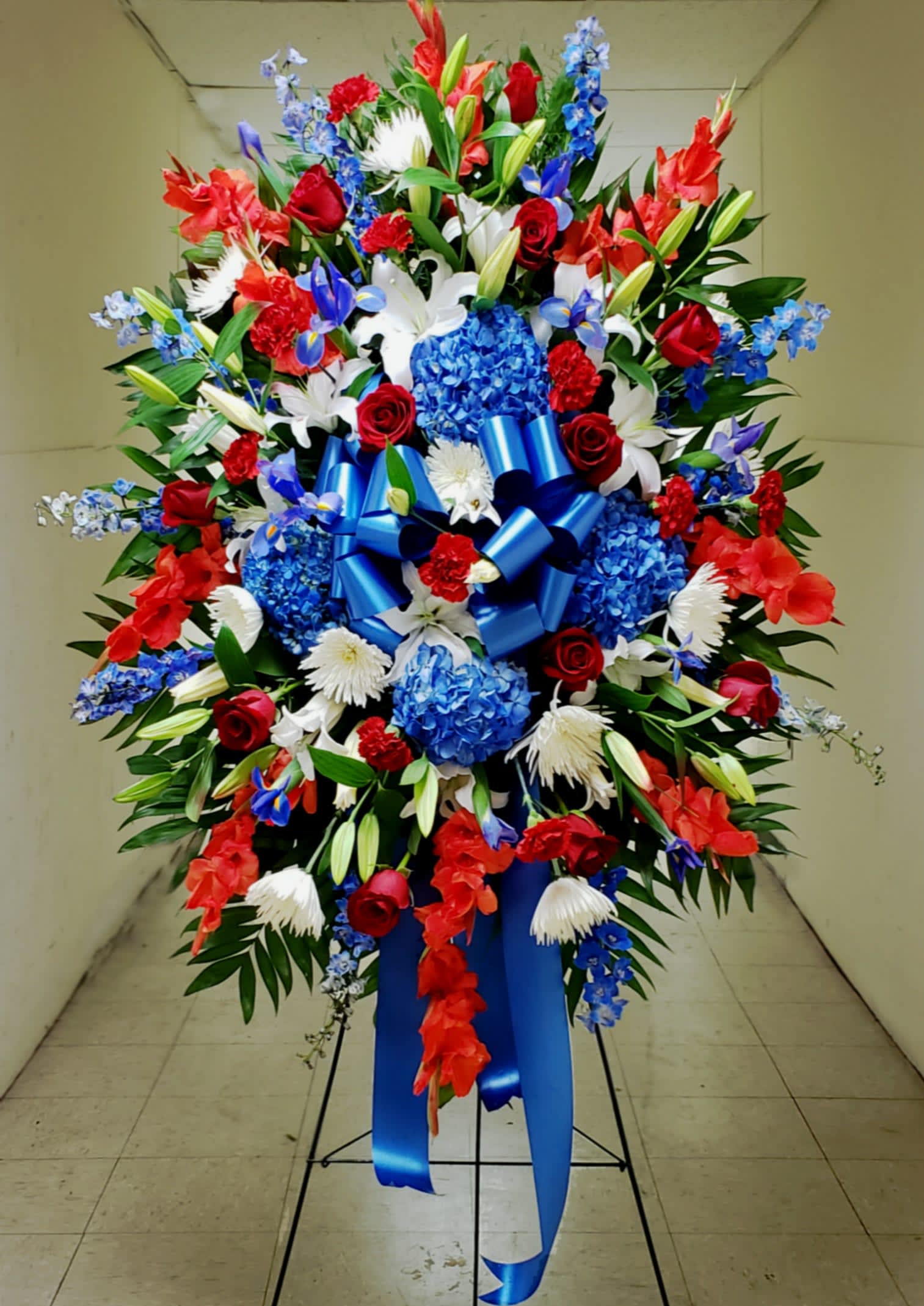 FULL HONORS STANDING SPRAY - OUR LARGEST AVAILABLE PATRIOTIC STANDING SPRAY COMES MOUNTED ON A TALLER 60" EASEL SPECIFICALLY INTENDED FOR FRONT-AND-CENTER  PLACEMENT AT MILITARY FUNERAL SERVICES AT ARLINGTON NATIONAL CEMETERY.  THIS EXCLUSIVE PREMIUM STANDING SPRAY MEASURES OVER 6' TALL, TOP STEM TO BOTTOM STEM, AND CONSISTS OF A DOZEN PREMIUM FREEDOM RED  ROSES, WHITE HYBRID ORIENTAL LILIES, NATURAL DEEP BLUE HYDRANGEAS, NATURAL BLUE SEA WALTZ DELPHINIUM, SELECT GRADE RED CARNATIONS, DOMESTICALLY GROWN GLADIOLAS, WHITE FUJI CHRYSANTHEMUMS, AND BLUE IRISES.  THE BOW IS MADE WITH A WEATHERPROOF SATIN MATERIAL.  A PRINTED SASH CAN BE ADDED FOR $15.00.  AFTER THE SERVICE, THE SPRAY IS EASILY REMOVED FROM ITS EASEL AND IS DESIGNED TO LAY FLAT ON THE GROUND AS A GRAVE BLANKET. 