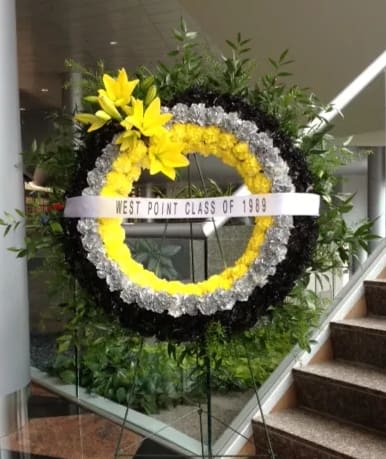 UNITED STATES MILITARY ACADEMY WEST POINT WREATH - CUSTOM 36" DIAMETER WREATH ON EASEL IN UNITED STATES MILITARY ACADEMY WEST POINT COLORS.  BLACK, GOLD (YELLOW), AND GRAY FRESH FLORAL WREATH FLOWERS IN A WET FLORAL FOAM WITH SALAL, ITALIAN RUSCUS, AND A SPRAY OF YELLOW LILIES.  A SINGLE HORIZONTAL PRINTED BANNER IS INCLUDED.