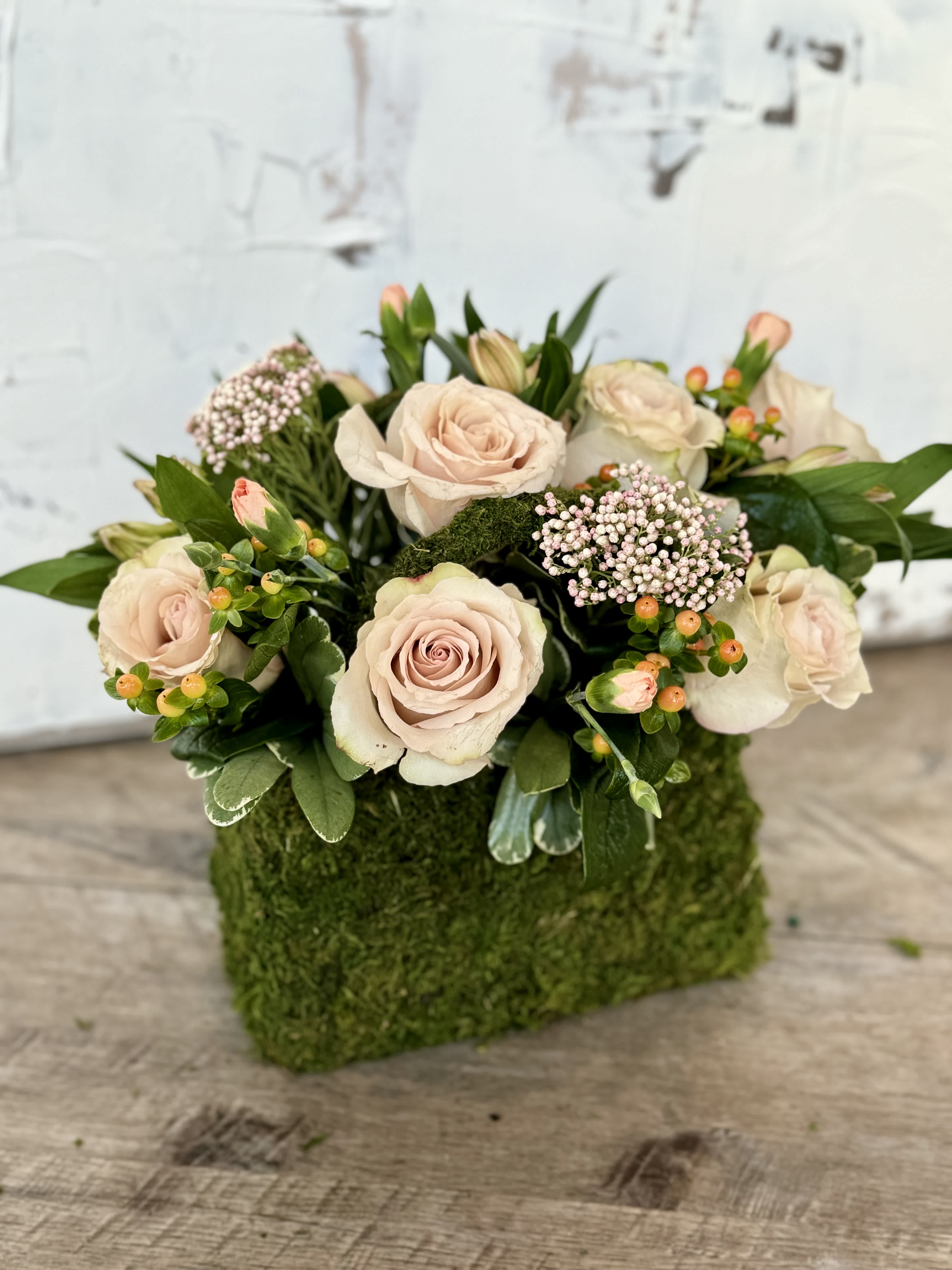 Blushing Blooms  - Crafted within a charming moss purse, this floral arrangement exudes grace and sophistication.  Delicate light pink roses intertwine with the subtle beauty of pink rice flower, while the hypericum berries add a touch of depth and richness.  