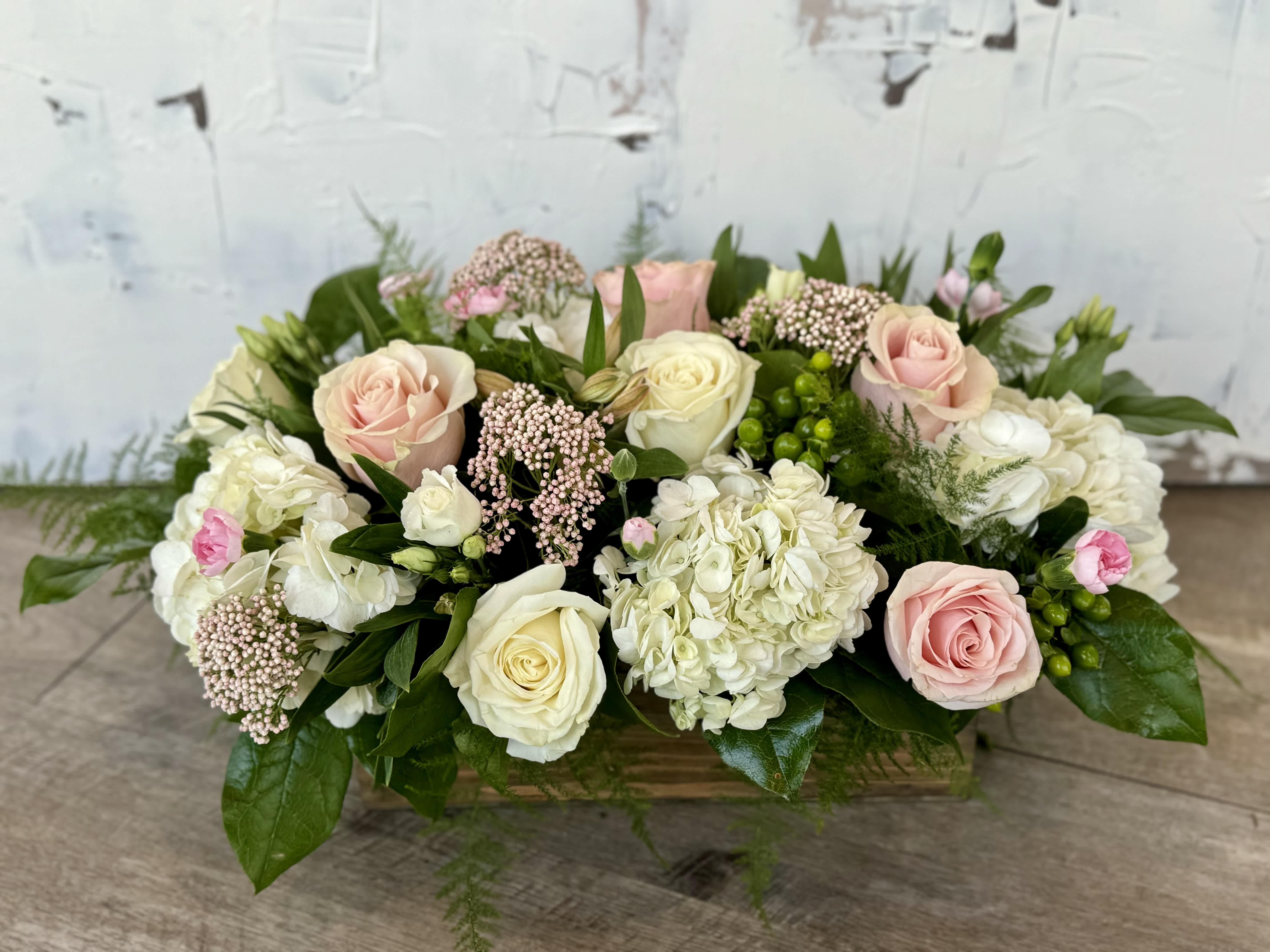 Graceful Blush  - Experience the timeless elegance of nature with this enchanting floral arrangement nestled in a rustic wooden box.  Delicate light pink roses and pink wax flower intertwine gracefully, while the purity of white hydrangea blooms adds a touch of sophistication.  Vibrant green hypericum berries provide a delightful contrast, enhancing the arrangements natural charm.  
