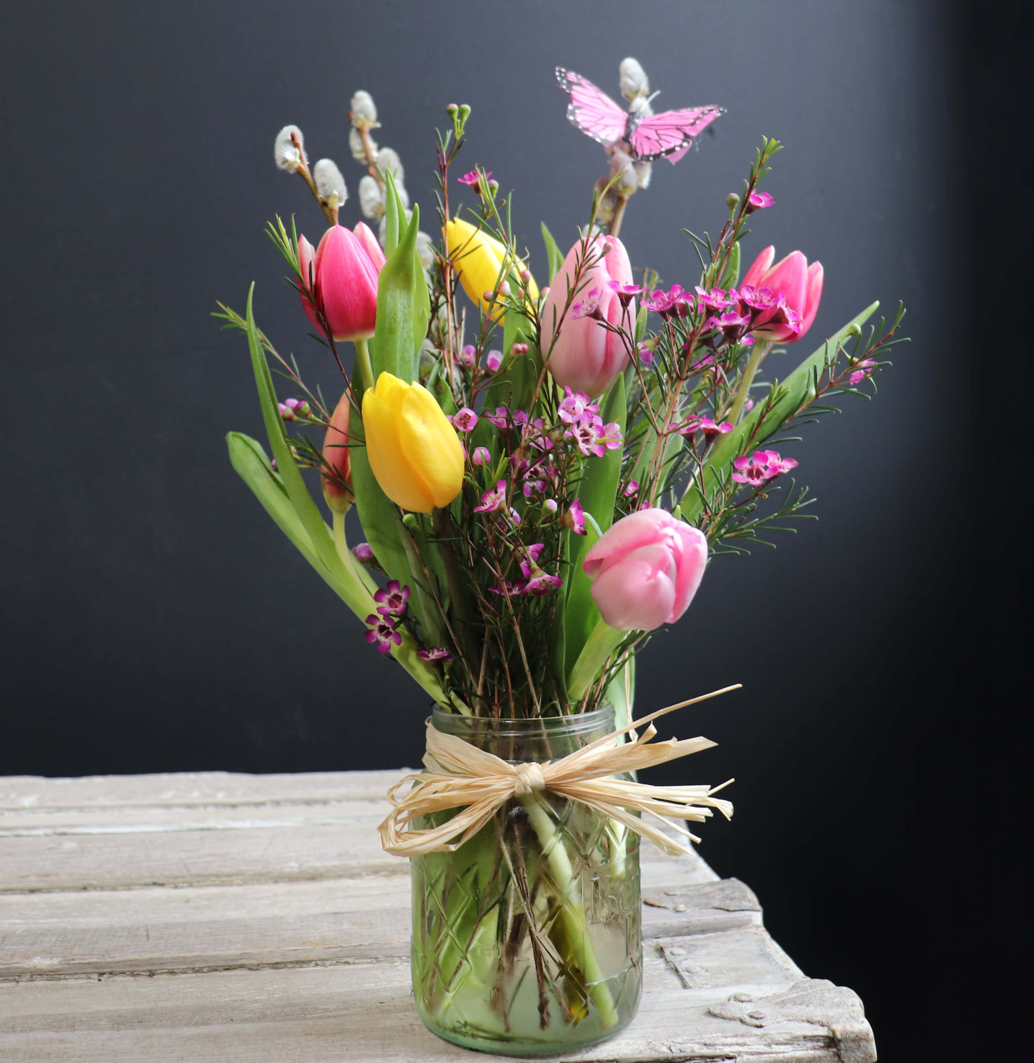 Tulip Melody - A fun bright colorful arrangement full of spring color and life! With a variety of colored tulips in a pint mason jar (for standard - approximately 10" tall) (premium size quart mason jar) with a butterfly to top it off, this design definitely is a day brightener!