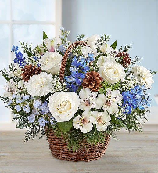 Winter Wishes Basket - With all the colors of winter, this beautiful arrangement would make the perfect addition to your Winter table. An incredible assortment of white roses, pinecones, lilies, and blue delphinium are arrangement in a wicker basket. This is the perfect centerpiece to make table extra special.  Your arrangement will be made to order. No two arrangements are identical. We try to recreate the picture, however it will not be exact.