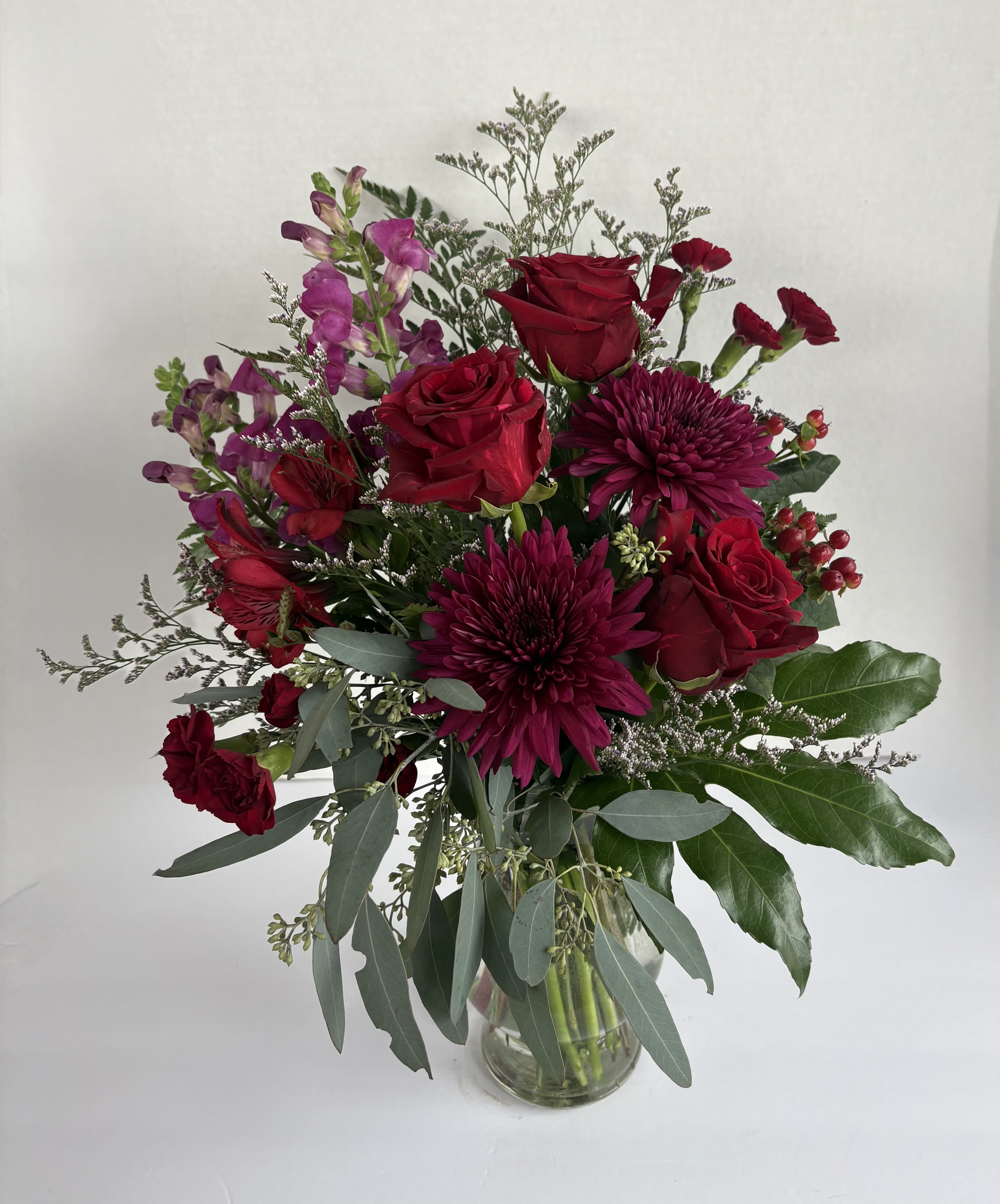 Warm Embrace  - This stunning arrangement is made up of red roses, dark purple cremones, red mini carnations, alstromeria, hypericum, purple snapdragons, and caspia, accented with seeded eucalyptus and fatsia leaf. The perfect gift to acknowledge a loved one. Say "I love you" with a Warm Embrace.