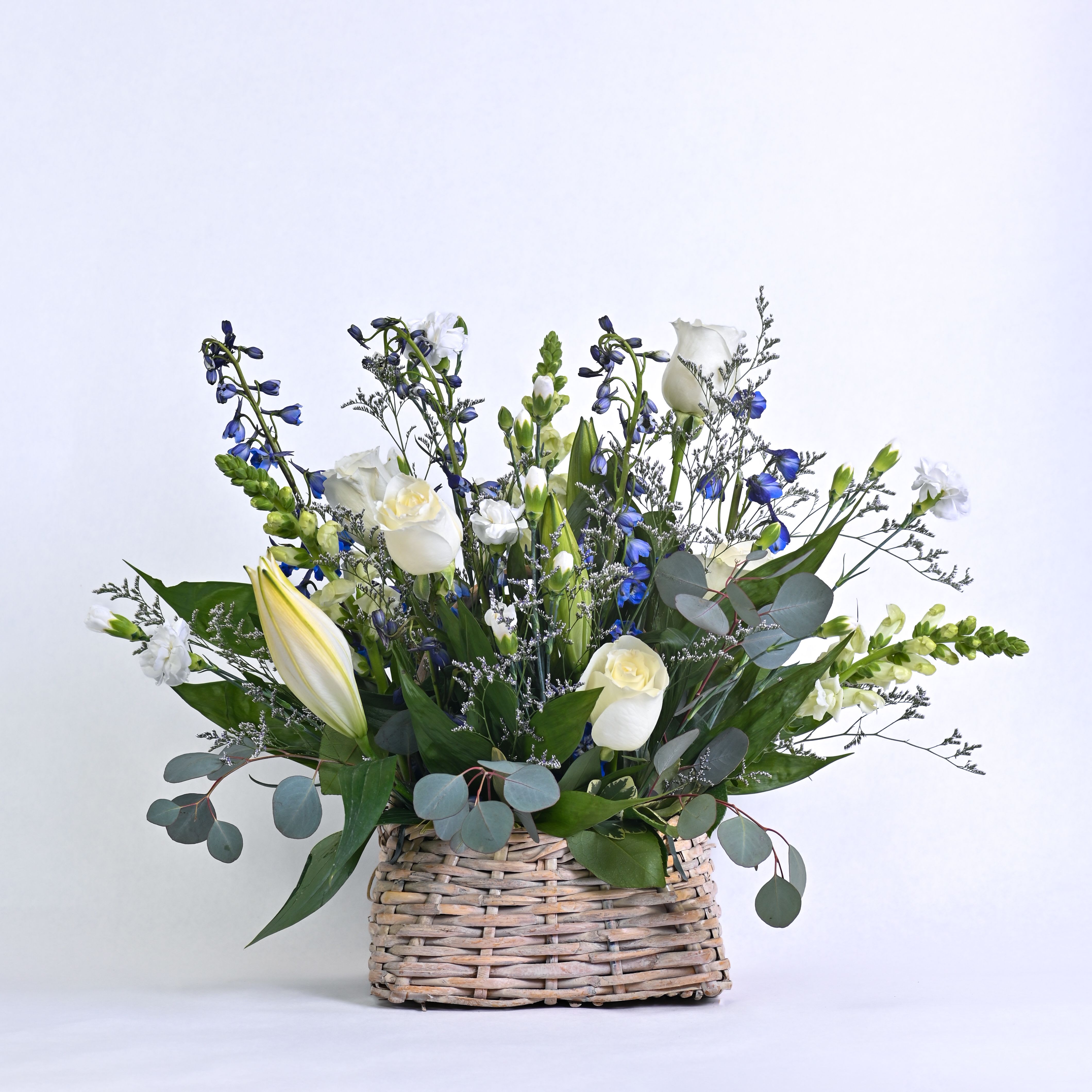 Peaceful Wishes - Tranquility is the word that immediately comes to the forefront in describing this serene yet exceptional floral creation of white lilies,  blue delphinium, white roses, white mini carnations and white snapdragons in a willow basket.