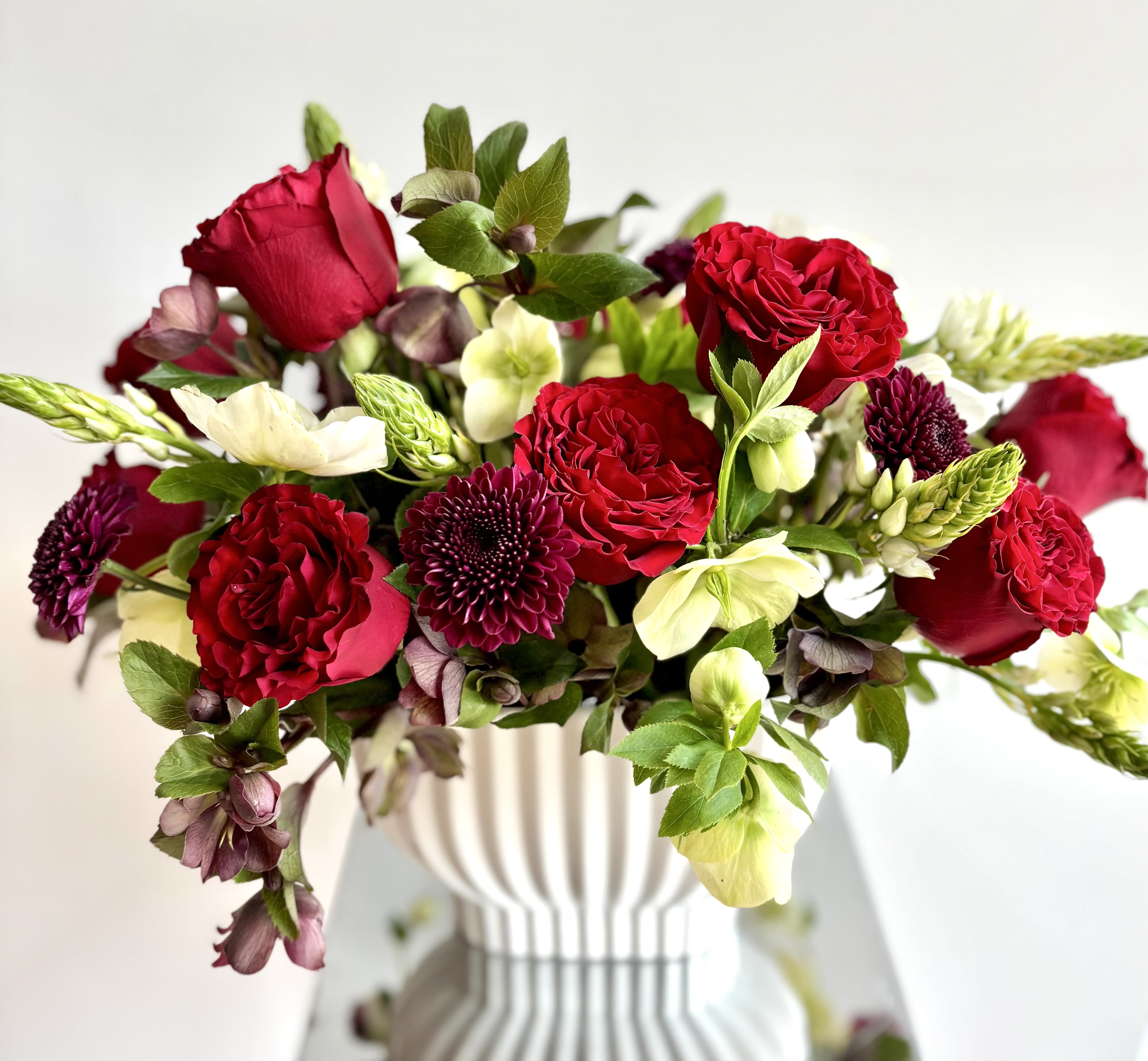 L'Amour - Stunning lush flower arrangement with premium Red Heart roses, hellebore, cremons and white Star of Bethlehem as focal flowers arranged with additional flowers in a red/burgundy-white-green color palette in a rippled ceramic vase.