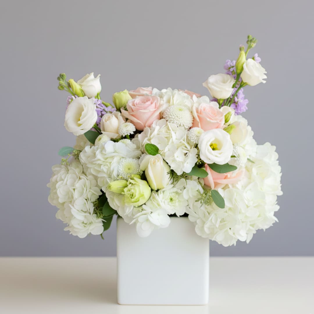 Garden Whisper - Send the Garden Whisper Arrangement Send a breath of elegance with our Garden Whisper arrangement! This sophisticated and airy design showcases a serene palette of blush pinks and creamy whites, perfect for expressing heartfelt sentiments, celebrating an anniversary, or bringing a touch of high-end garden beauty into a Southwest Florida home.  This graceful display features cloud-like white hydrangeas, romantic blush roses, and fragrant pastel stock flowers that provide the height and texture you adore. We’ve elevated this signature look with the delicate, ruffled petals of premium white lisianthus, replacing standard greens for a truly luminous finish. All these exquisite blooms are artfully nestled in a 5" sleek white ceramic cube, creating a clean, modern, and captivating presentation.  Let us help you share a moment of refined beauty. Order the Garden Whisper today and let your message bloom with style!