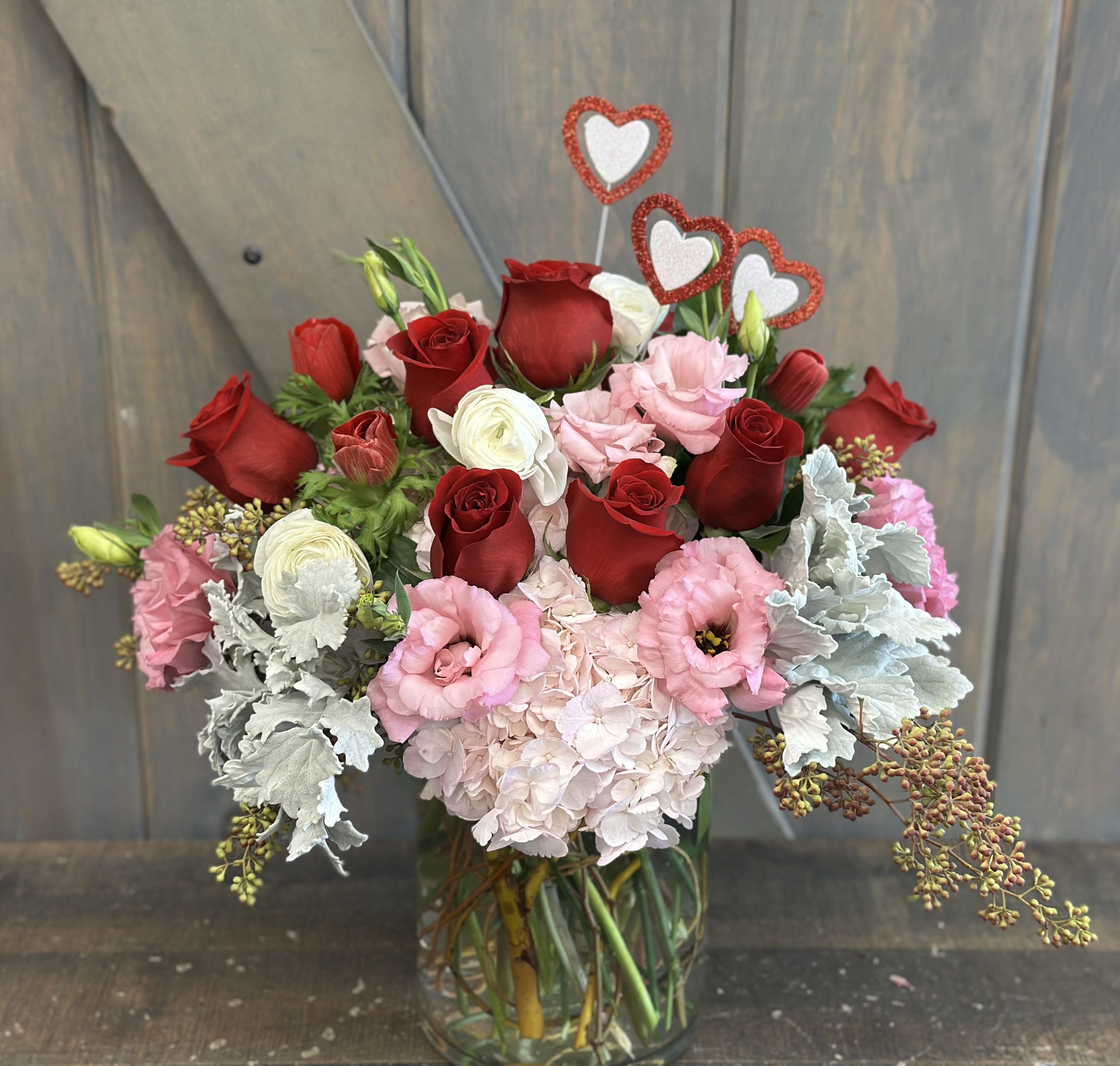 Shirley Temple - This youthly romantic arrangement is bound to put a smile on your recipients face. Flowers included in this design are Hydrangea, Roses, Lisianthus, Ranunculus, Anemones, Dusty Miller, and seeded eucalyptus all within a glass cylinder vase with hearts Included