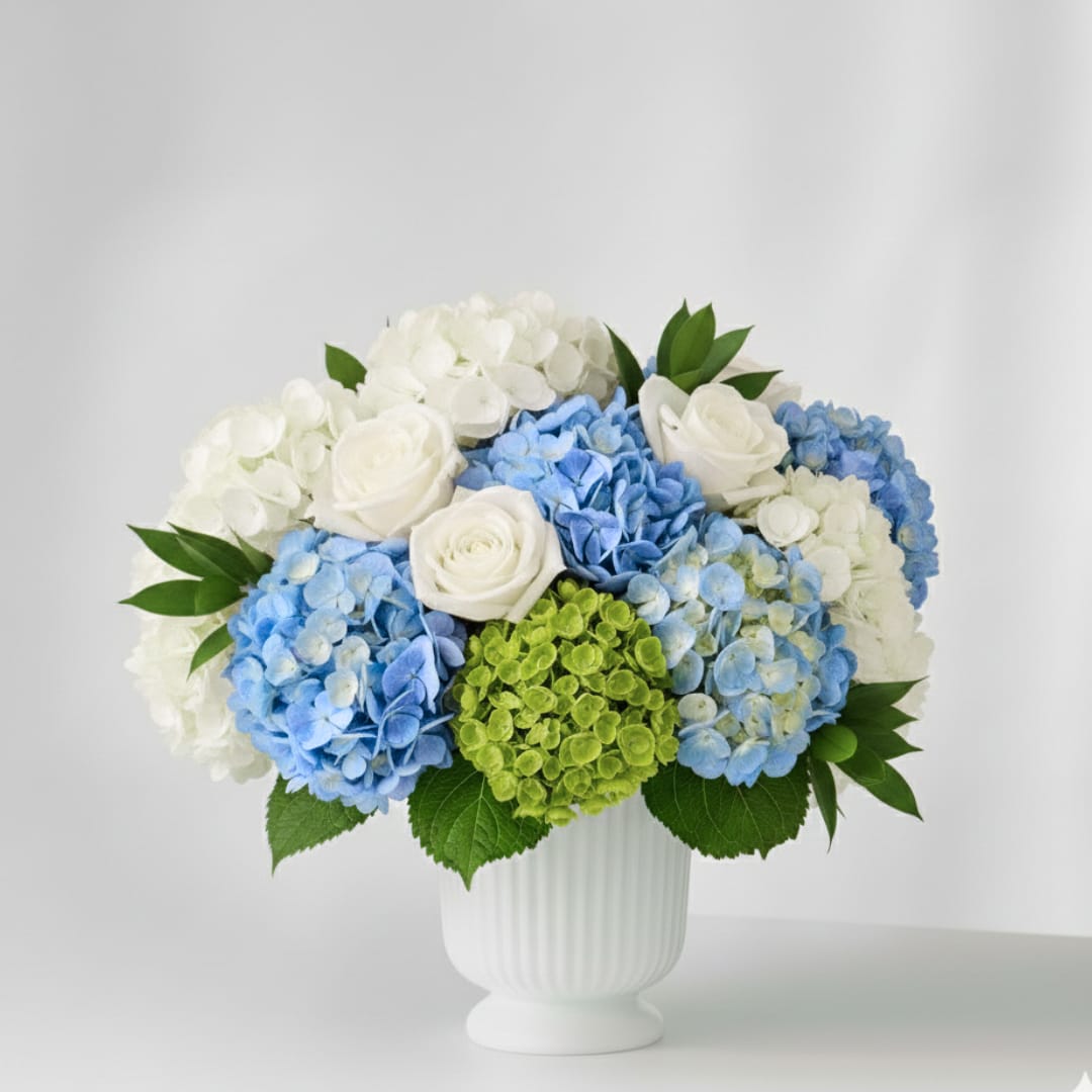 Coastal Elegance - Coastal Elegance  Inspired by the breezy, sophisticated charm of the Gulf Coast, Coastal Elegance is a signature Estero Market Florist design that balances upscale refinement with an organic, whimsical spirit.  This lush arrangement features premium White Hydrangeas and cloud-like Blue Hydrangeas, creating a soft, textural foundation for our favorite high-petal-count White Roses. Tucked within the blooms are stems of Israeli Ruscus, adding a touch of verdant, garden-fresh movement. Presented in a timeless, fluted white ceramic urn, this piece is designed to feel like a breath of fresh air on a sun-drenched veranda.  Aesthetic: Airy, Whimsical, and Upscale  Key Florals: Premium White Roses, Blue and White Hydrangeas, and Israeli Ruscus  Vase: Classic White Fluted Ceramic Urn  Perfect For: Coastal home decor, elegant birthdays, or a sophisticated "just because" gift.