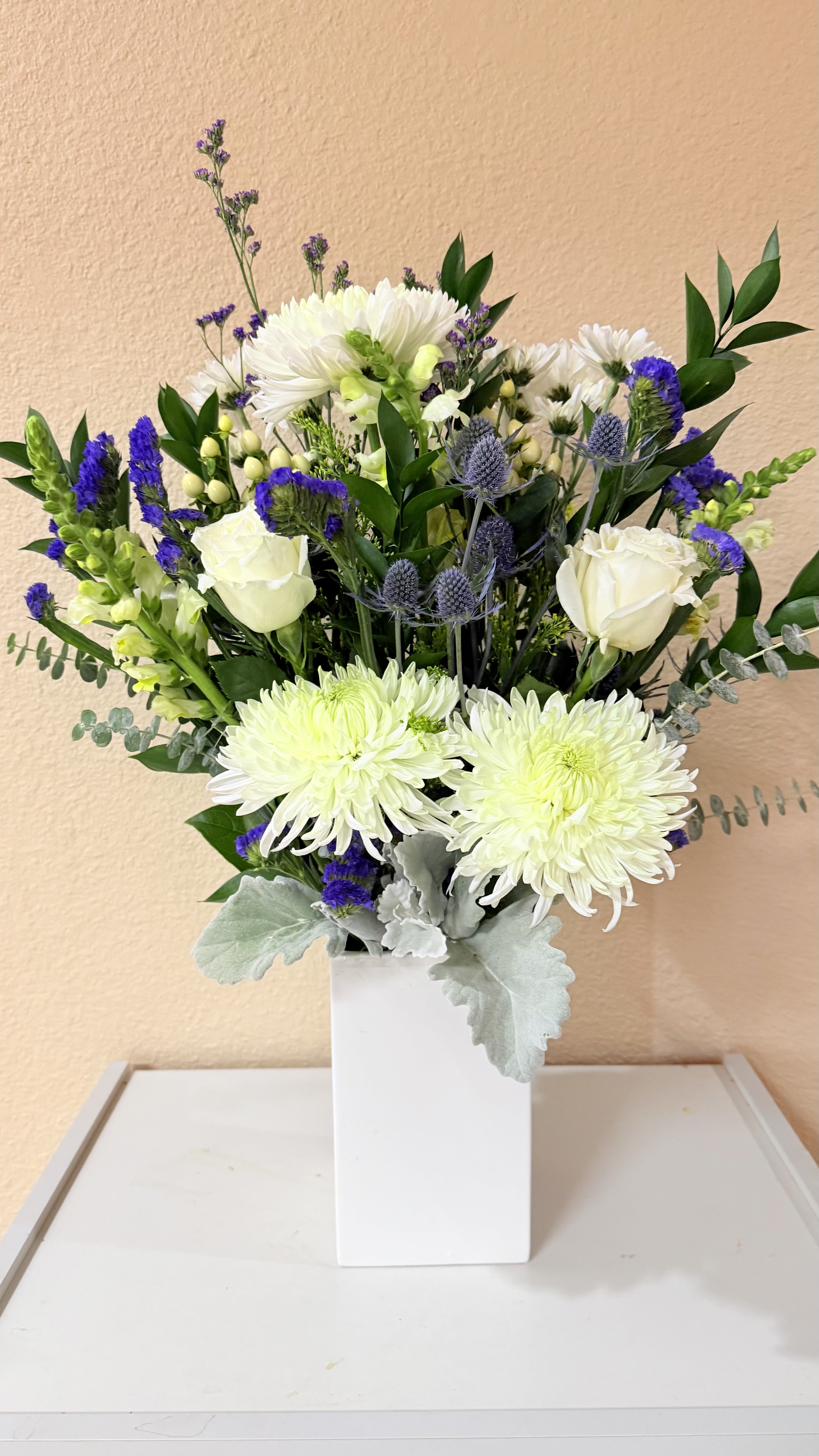 Winter Serenity  - A calming blend of soft whites, cool greens, and soothing shades of blue and lavender. features elegant white roses, crisp mums, delicate snapdragons, blue thistle, limonium , dusty miller, and lush greenery all arranged in a modern white ceramic vase.