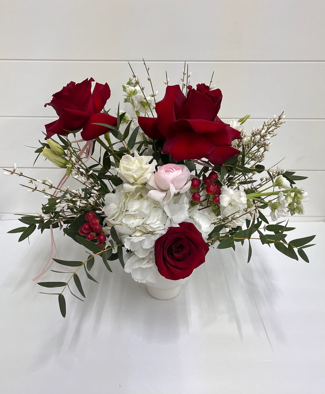 Love In Bloom - Love in Bloom is a timeless expression of romance, featuring reflexed red roses at the heart of the design, softened by lush white hydrangea and delicate light pink ranunculus. Finished with airy filler and styled in a cream compote bowl, this arrangement is a graceful celebration of love in full bloom.