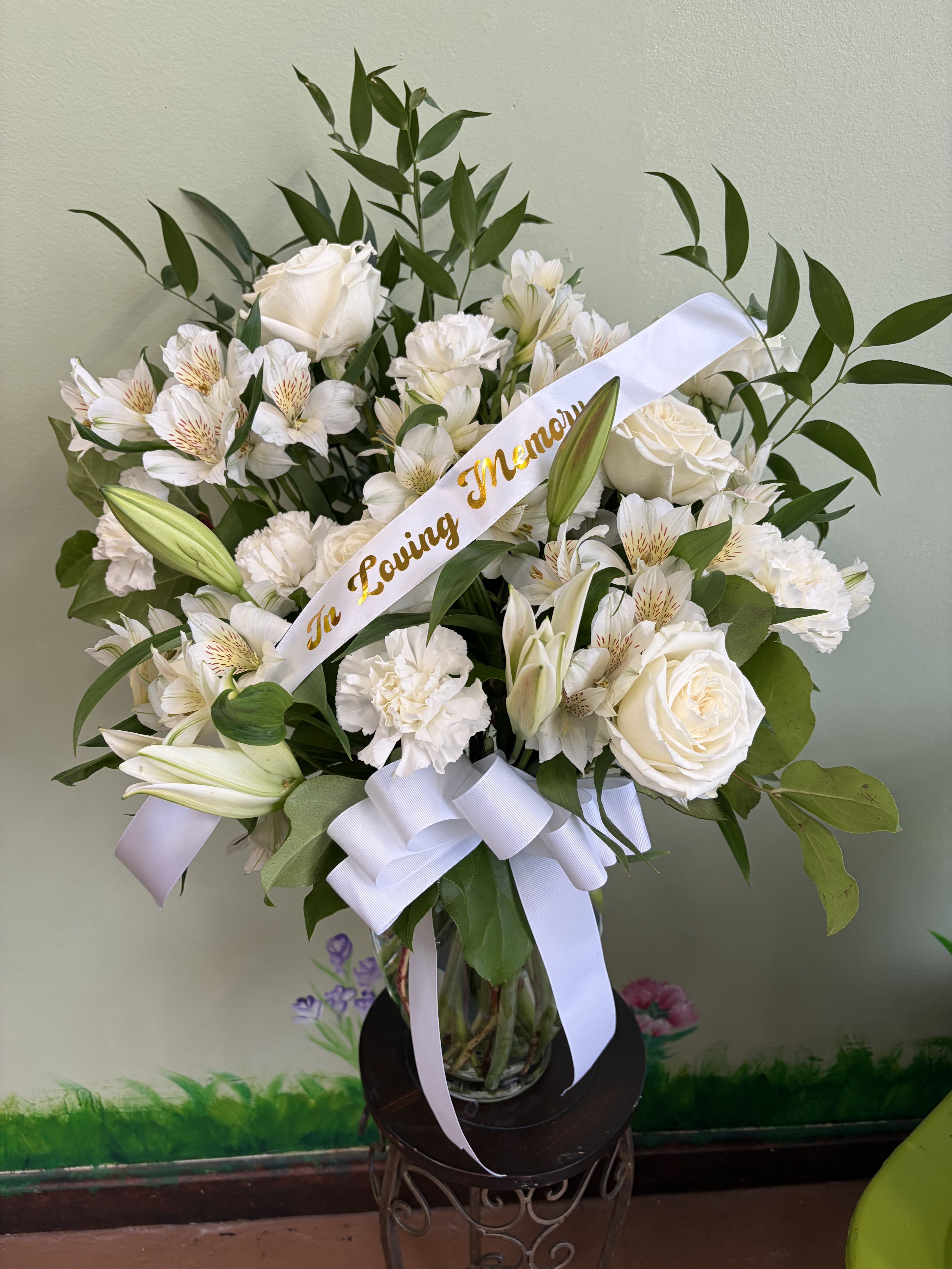 Pure Remembrance Vase w/ bow and ribbon - An elegant all-white arrangement featuring roses, lilies, carnations, and alstroemeria, accented with fresh green foliage. Designed to express peace, honor, and lasting remembrance, this vase arrangement offers a timeless and comforting tribute suitable for memorial services or home display.