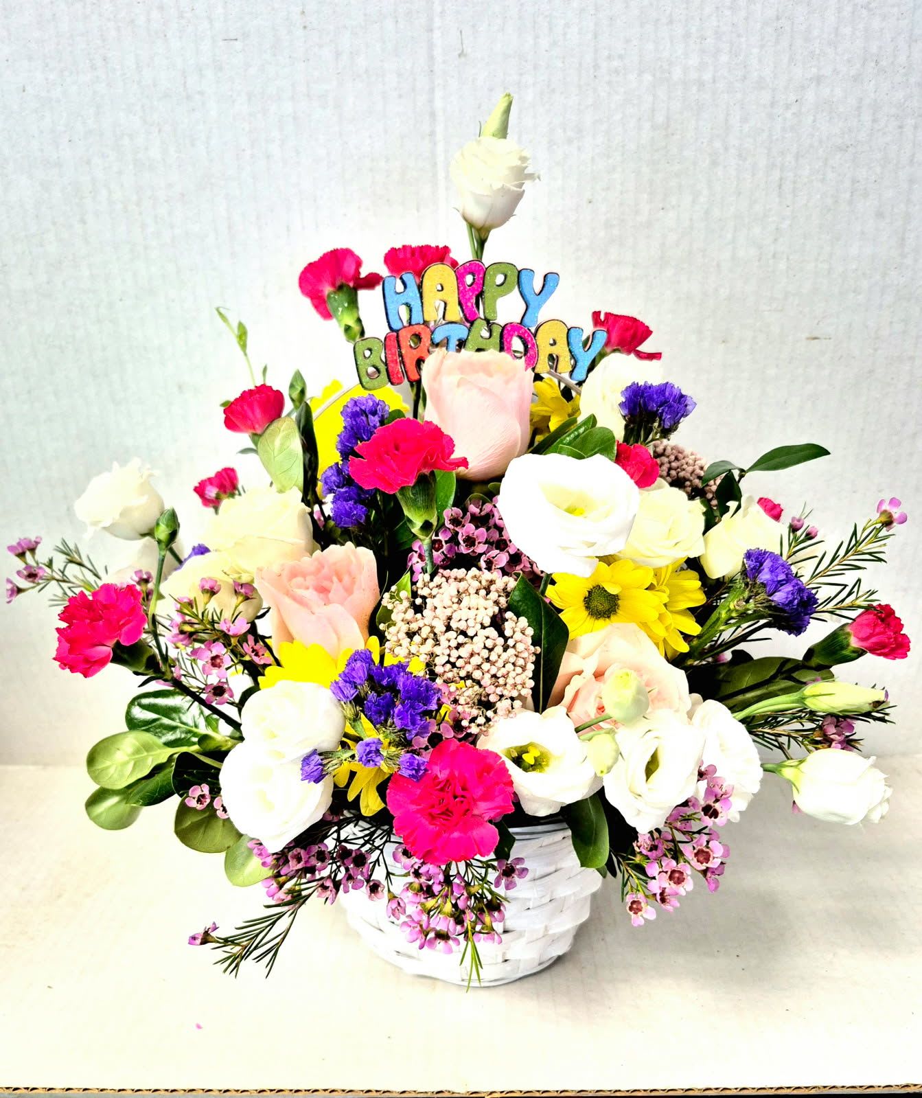 Birthday Deluxe Basket - An assortment of white and colorful flowers arranged in a white basket with a happy birthday pick. 