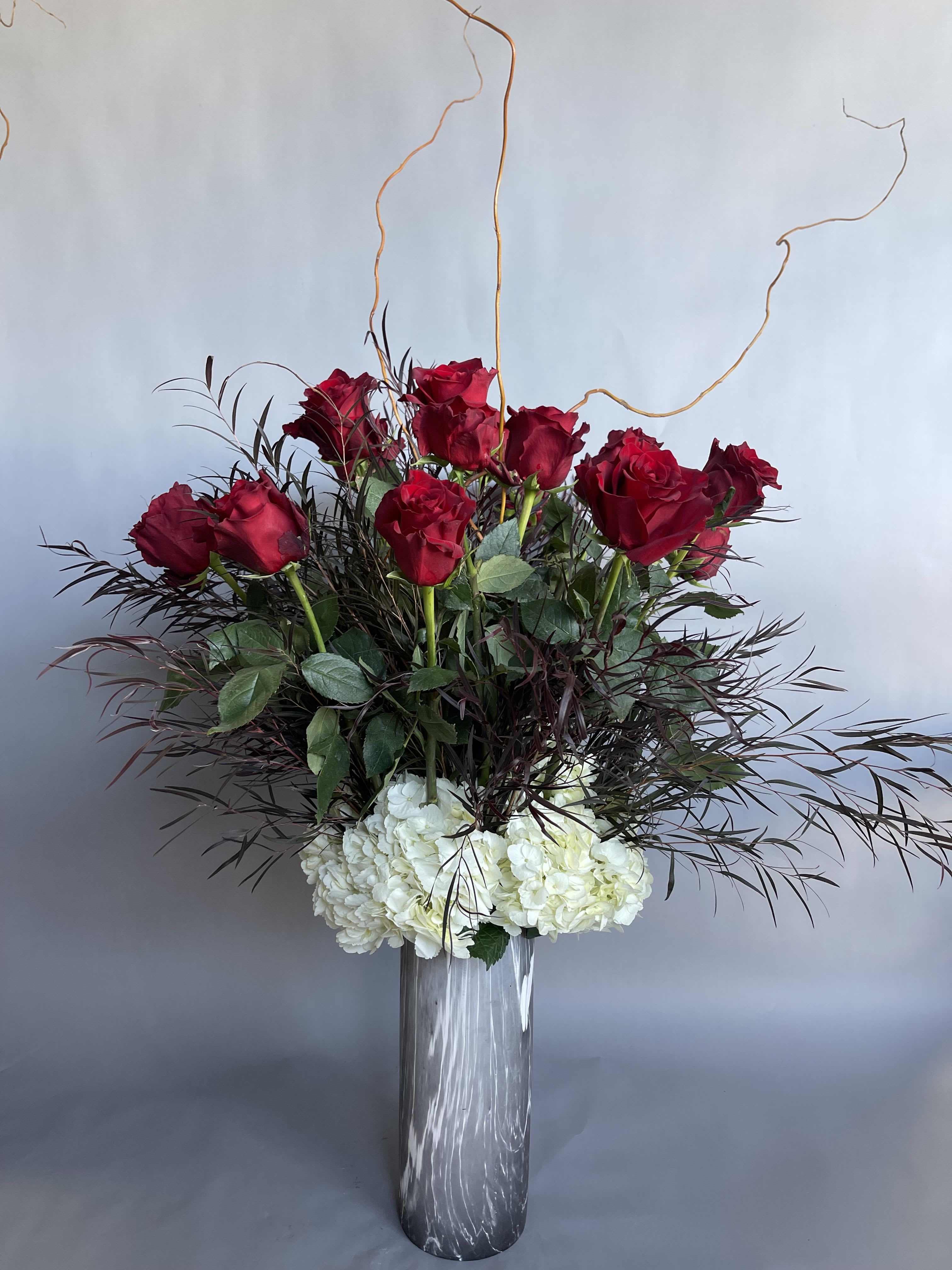 A lovely romance. A dozen and more. - * Stunning tall vase filled with white hydrangeas, a dozen long red roses and romantic dark filler. 