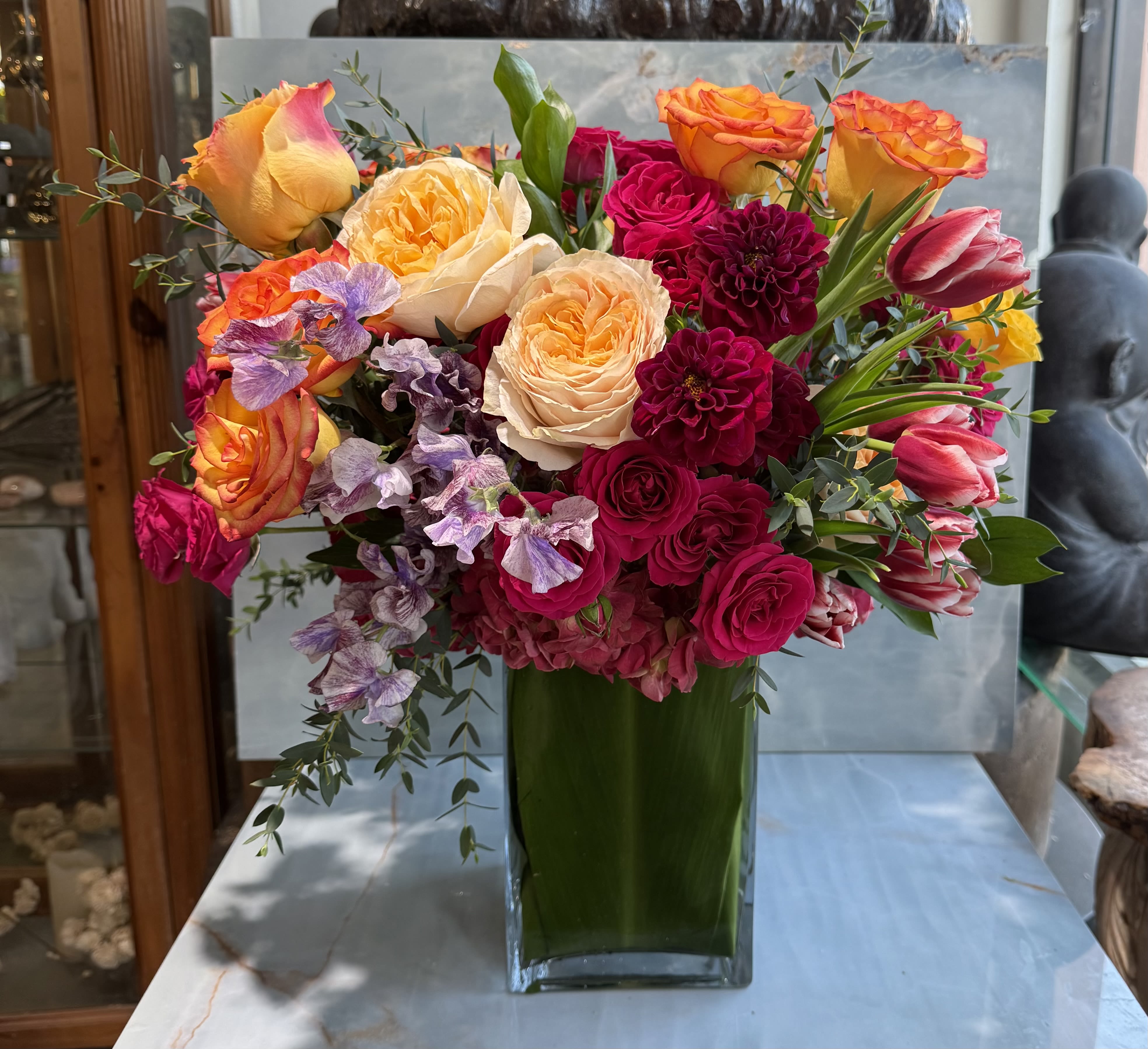 Sunset Sonata - This arrangement is lush, garden-inspired, and joyfully expressive. It features a rich mix of warm sunset roses in peach, apricot, coral, and golden yellow, paired with deep raspberry and burgundy booms that add depth and drama. Dahlias and tulips bring texture and movement, while soft lavender sweet peas and airy eucalyptus soften the palette and give it an organic, gathered-from-the-garden feel.