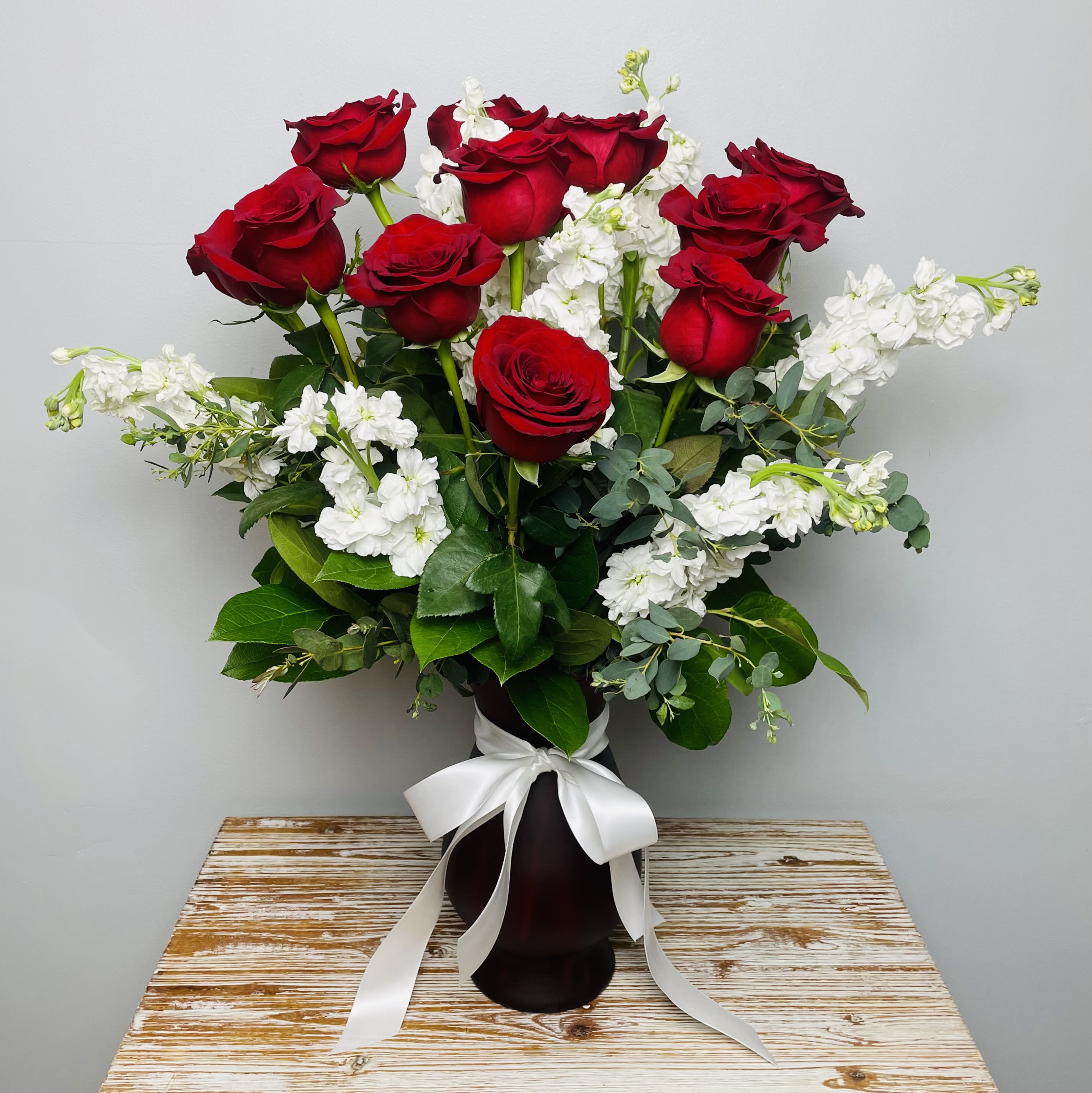 Luxe Red Roses - The luxurious take on a Dozen Red Roses. Twelve Long Stem Red Roses accented with fragrant White Stock and Eucalyptus in a Ruby Red Vase accented with a white silky soft satin ribbon.  Note: The Stock in this arrangement is fragrant. Stock gives off a sweet aromatic scent that many find as pleasant.  Approximately 24-26" Tall 