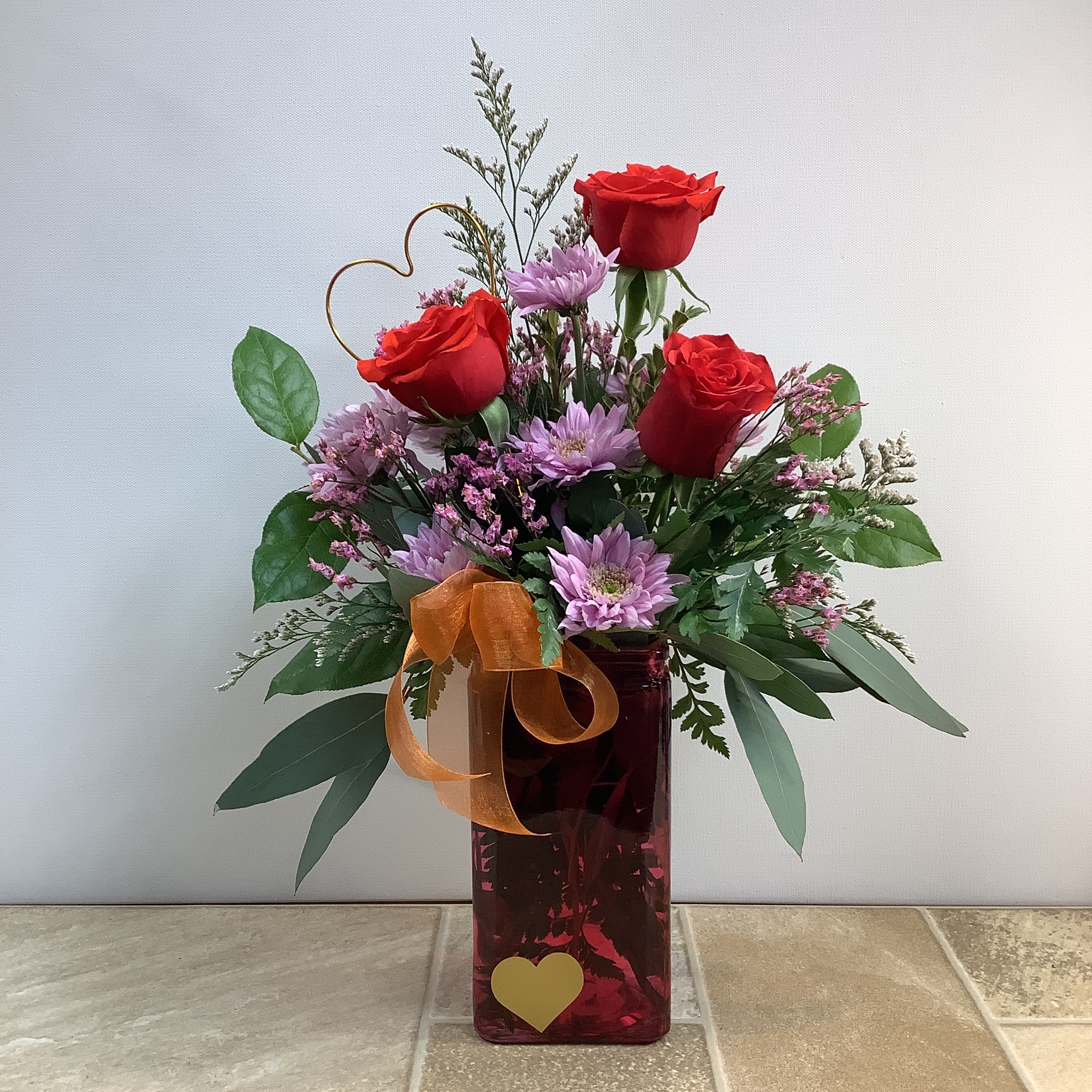 Love You Always - Send a little love with this unique bouquet of orange Nina roses, lavender daisies, and pink diamond limon, surrounded by a variety of green foliage.  A gold wire heart adds a special touch to the arrangement. The lovely blooms are arranged in a keepsake, dark pink glass vase with a pretty gold heart and a coordinating bow. Approximate size: 13” wide x 19” tall.  Primary flowers: rose, daisy