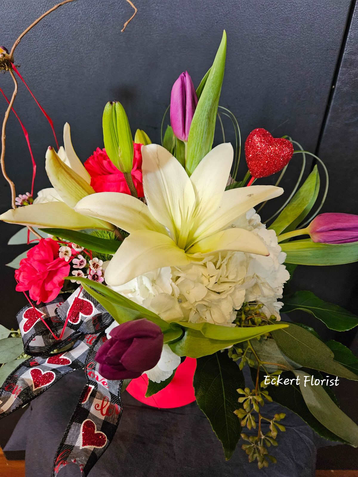 Eckert Florist's "Our Love Blooms" Bouquet *Local Delivery Only - Designed in a red cache pot, this round arrangement has a hint of spring throughout with tulips, hydrangea, lilies, carnations, filler flower, and designer greenery finished with a sparkling heart and designer ribbon. Flower colors, container and ribbon may vary. *Our local delivery only.