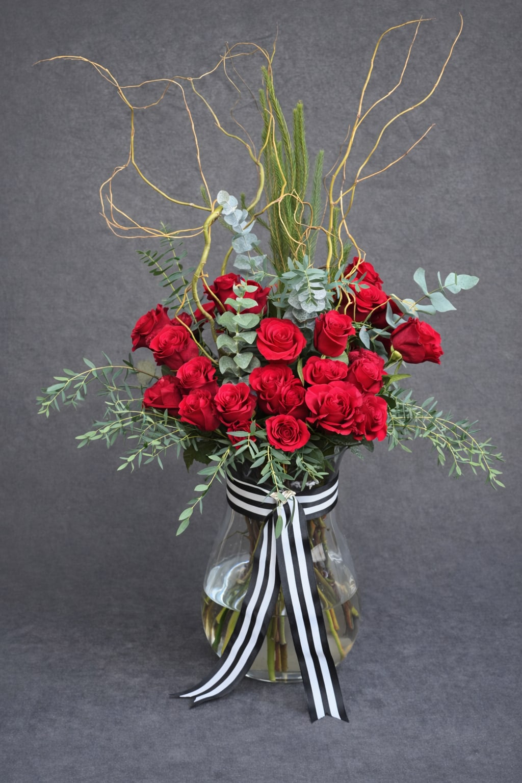 The Grand Gesture - Elegant long stemmed red roses, curly willow, fragrant eucalyptus, in a large vase tied with a black and white ribbon. Please note this is a large arrangement, vase is 18in in height roses are 60cm-70cm tall.    *Reasonable substitutions may be made due to floral availability. Will have the same overall look and feel. 