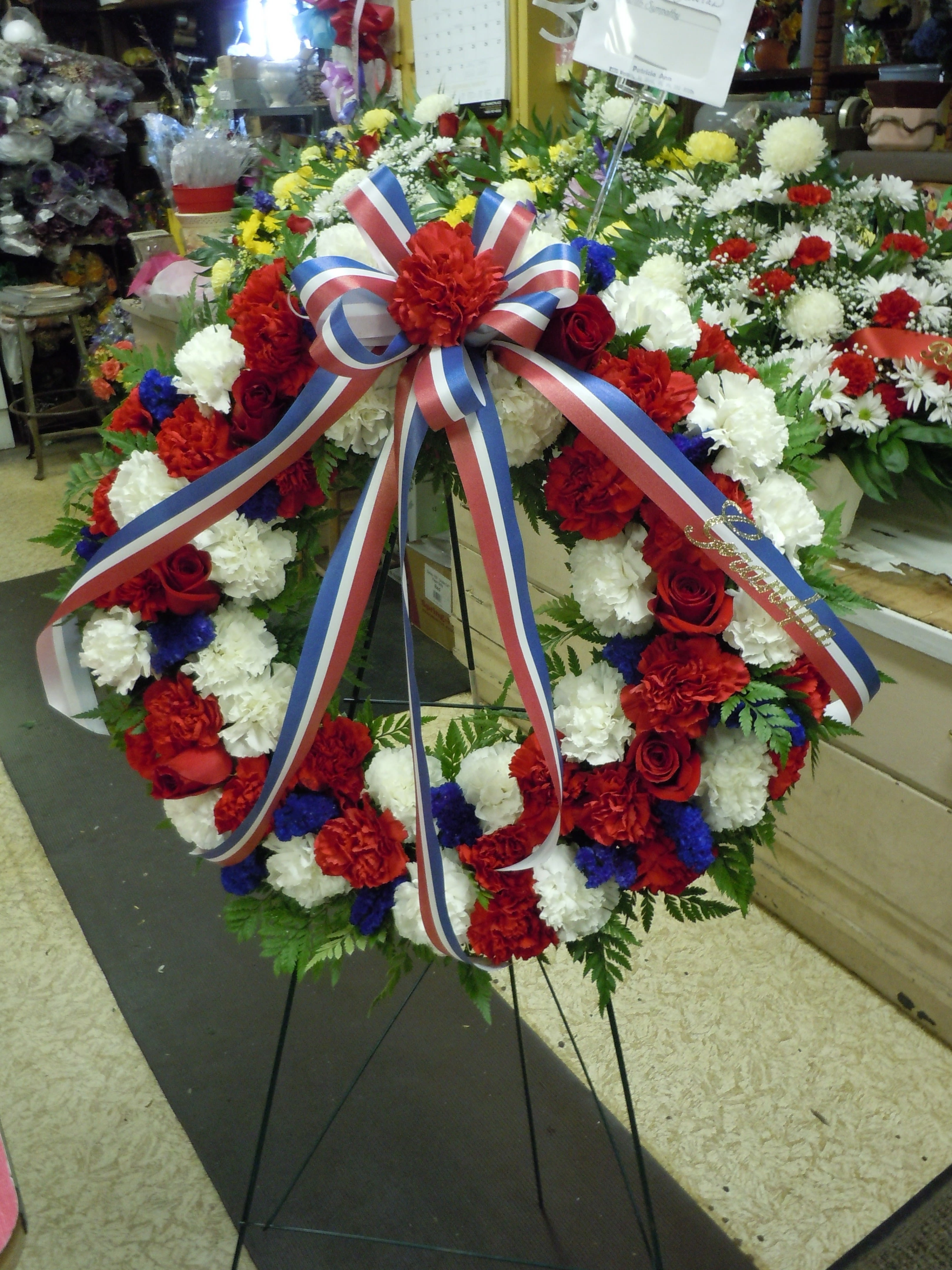 SP12 Standing Spray - A Round open wreath, includes red and white carnations, blue statice and red roses sprinkled, greens   Banner - $15.00 + extra  Substitutions may apply due to flower availability