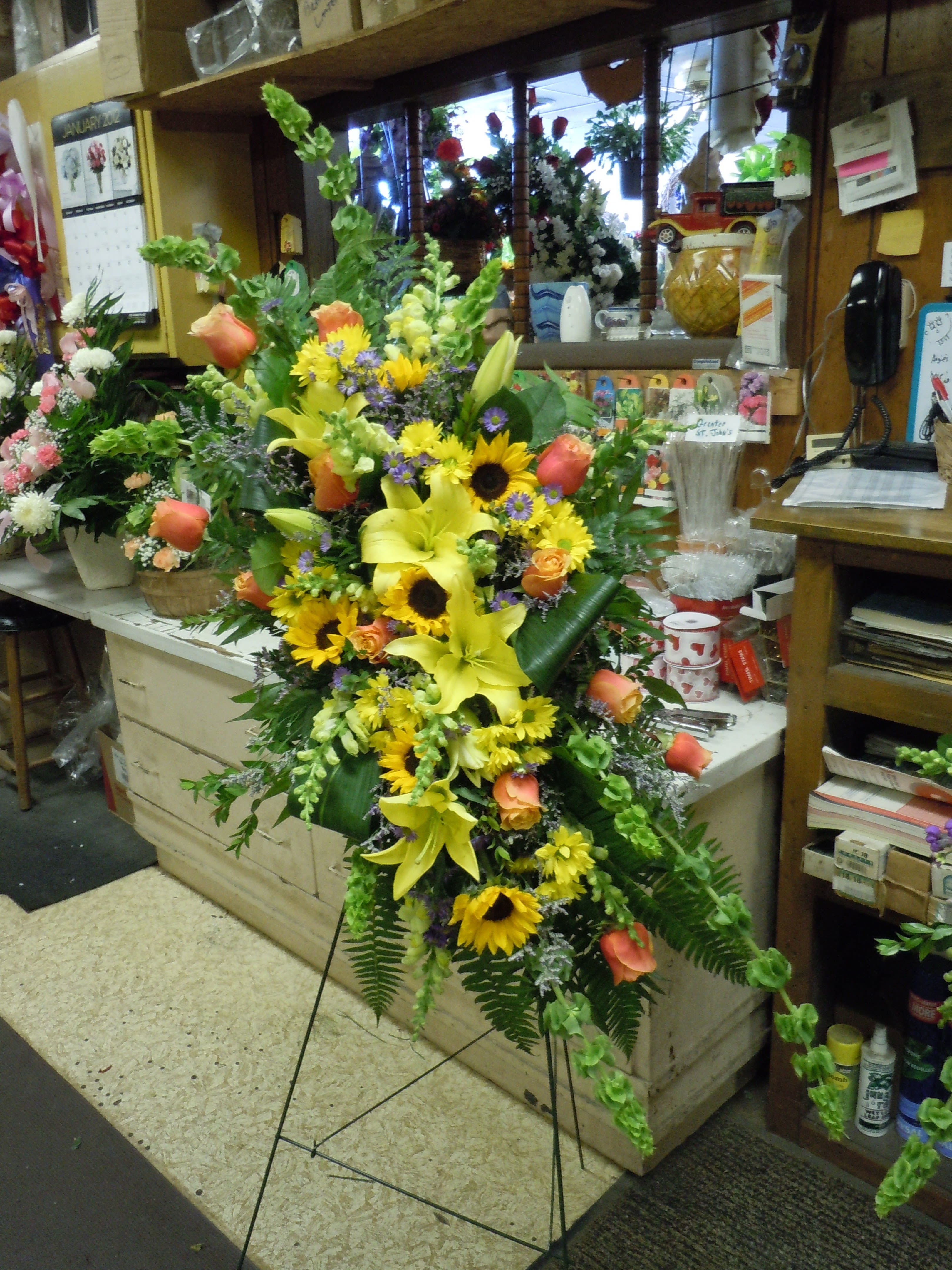 SP15 Standing Spray - A mix of Bells of Ireland, mini sunflowers, lilies, roses, snap dragons, greens and blue mist   Banner - $15.00 + extra  Substitutions may apply due to flower availability