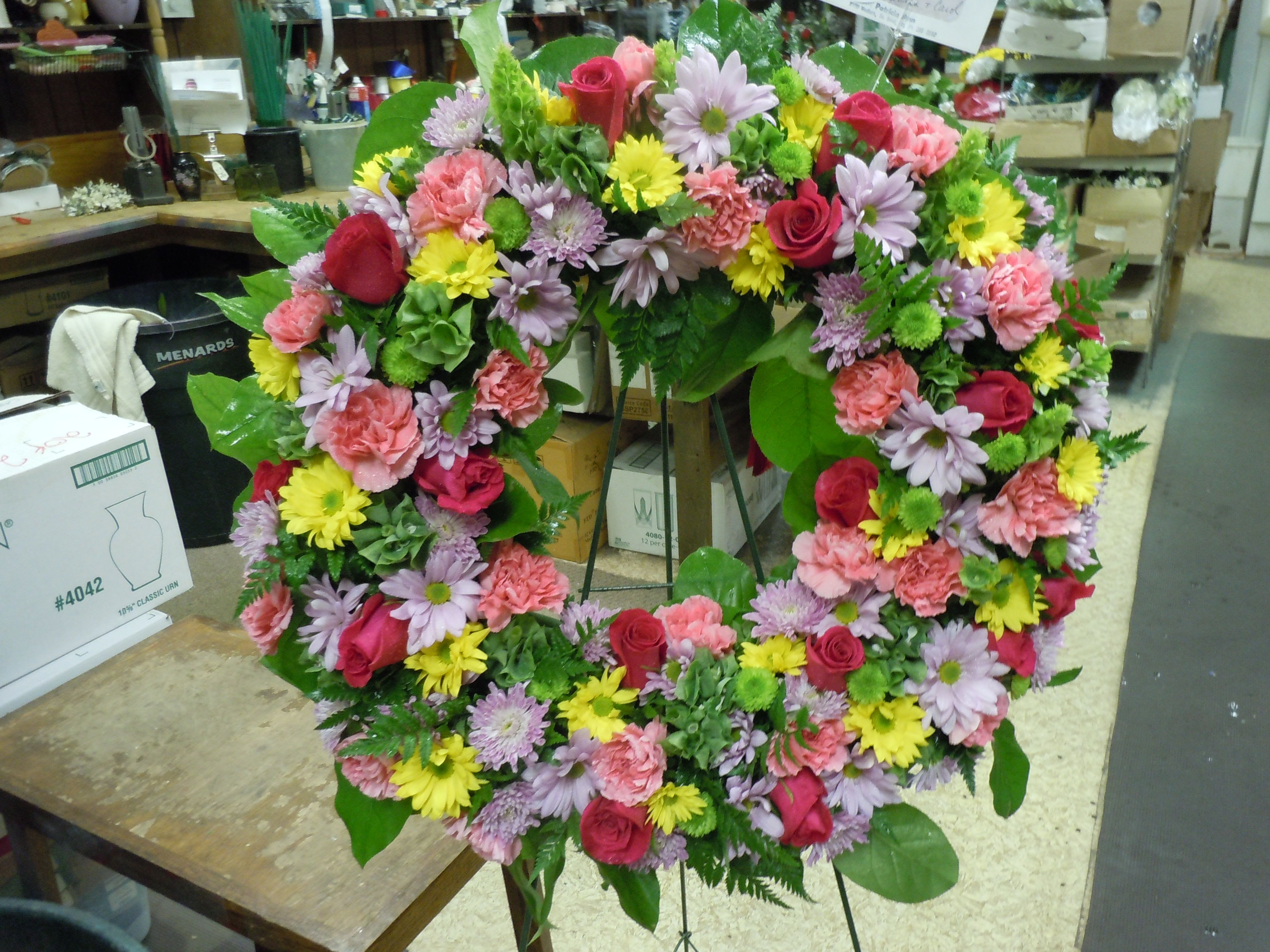 SP16 Standing Spray - An open wreath with a mix of roses, yellow daisies, Bells of Ireland, carnations, kermits, and greens.   Banner - $15.00 + extra   Substitutions may apply due to flower availability
