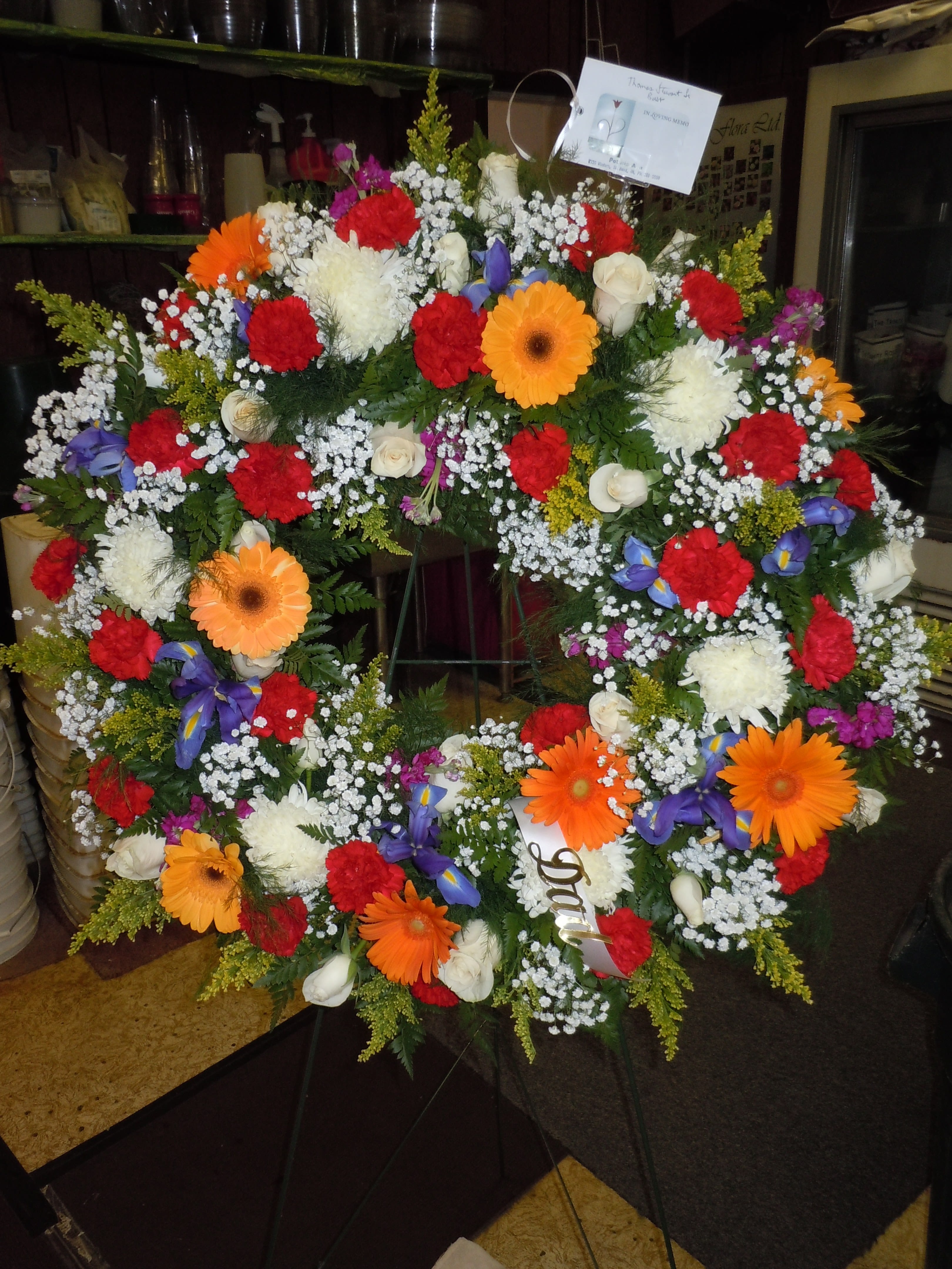 SP21 Standing Spray - An open wreath with a mixture of gerbera daisies, roses, fuji mums, iris, solidago, stock, baby's breath and greens.   Banner - $15.00 + extra  Substitutions may apply due to flower availability