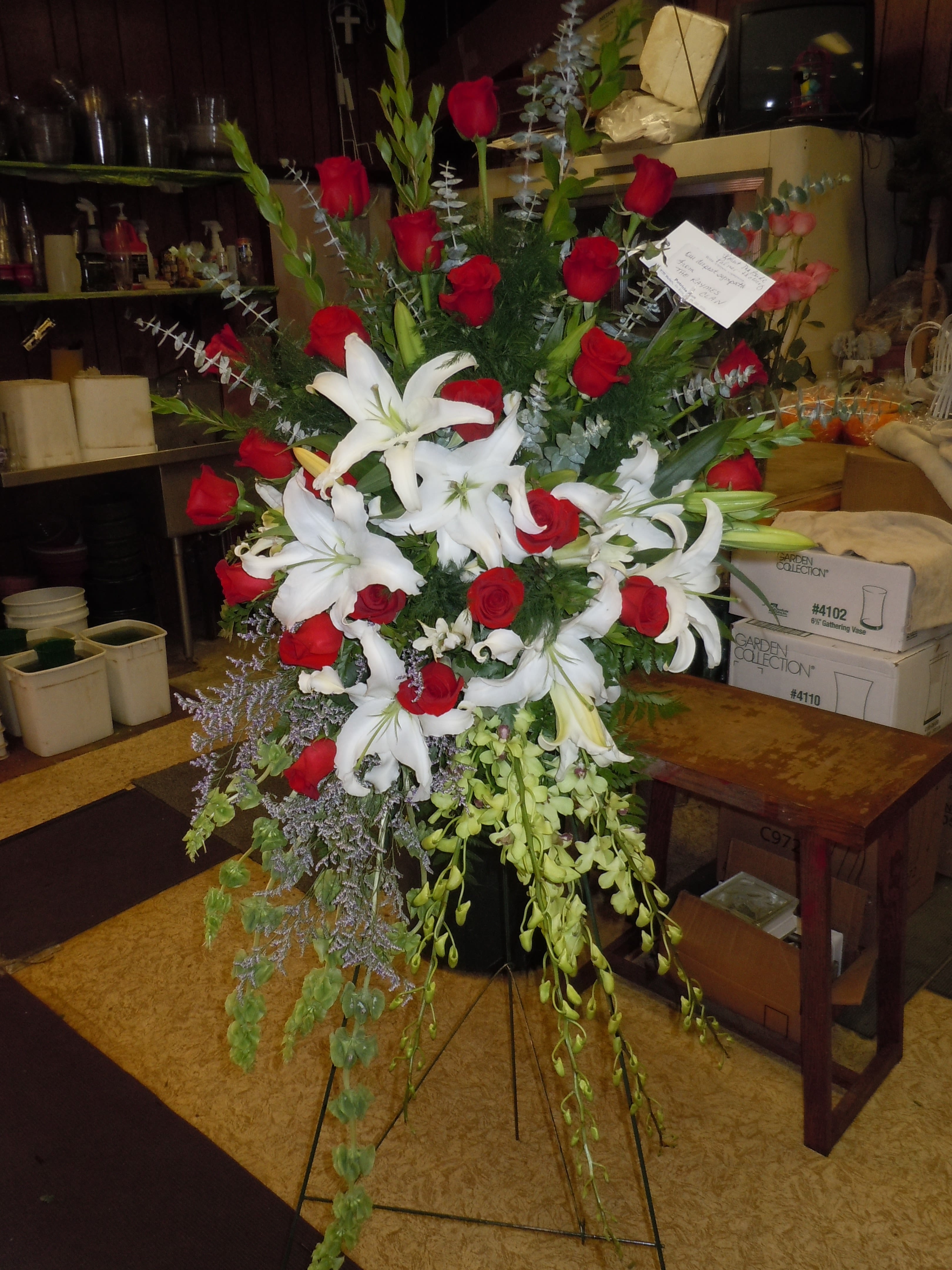 SP22 Standing Spray - A mixture of white lilies, red roses, bells of Ireland, dendro orchids, myrtle, blue mist and spiral euc and greens.   Banner - $15.00 + extra  Substitutions may apply due to flower availability