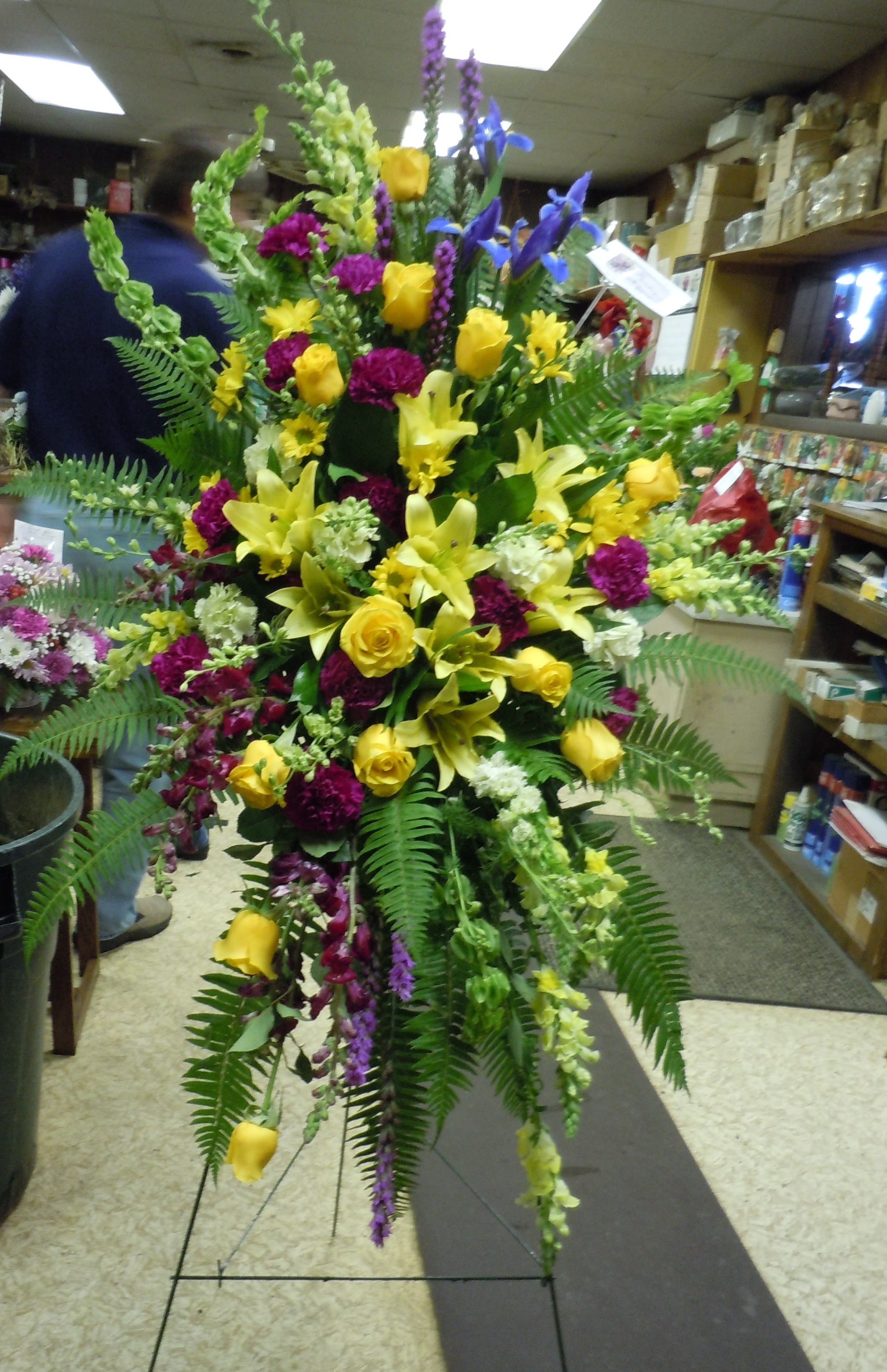 SP24 Standing Spray - A mixture of Bells of Ireland, dark fuscia carnations, yellow lilies, yellow snap dragons, sword fern, yellow roses, liatris, fuscia stock, yellow daisies, tree fern, leather leaf    Banner - $15.00 + extra  Substitutions may apply due to flower availability