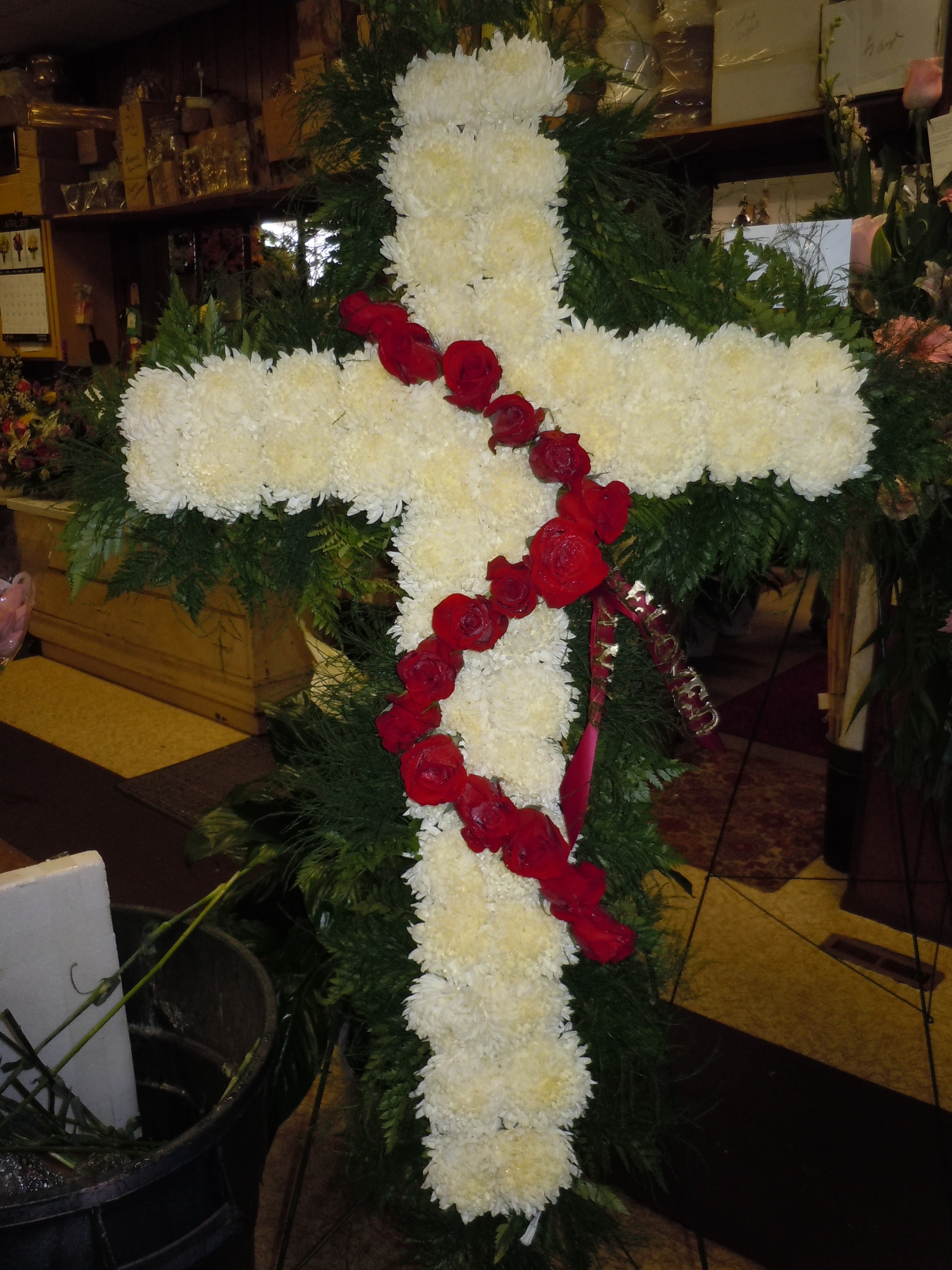 SP28 Standing Spray - A cross with a mixture of  roses, fuji mums,  and greens.   Banner - $15.00 + extra  Substitutions may apply due to flower availability