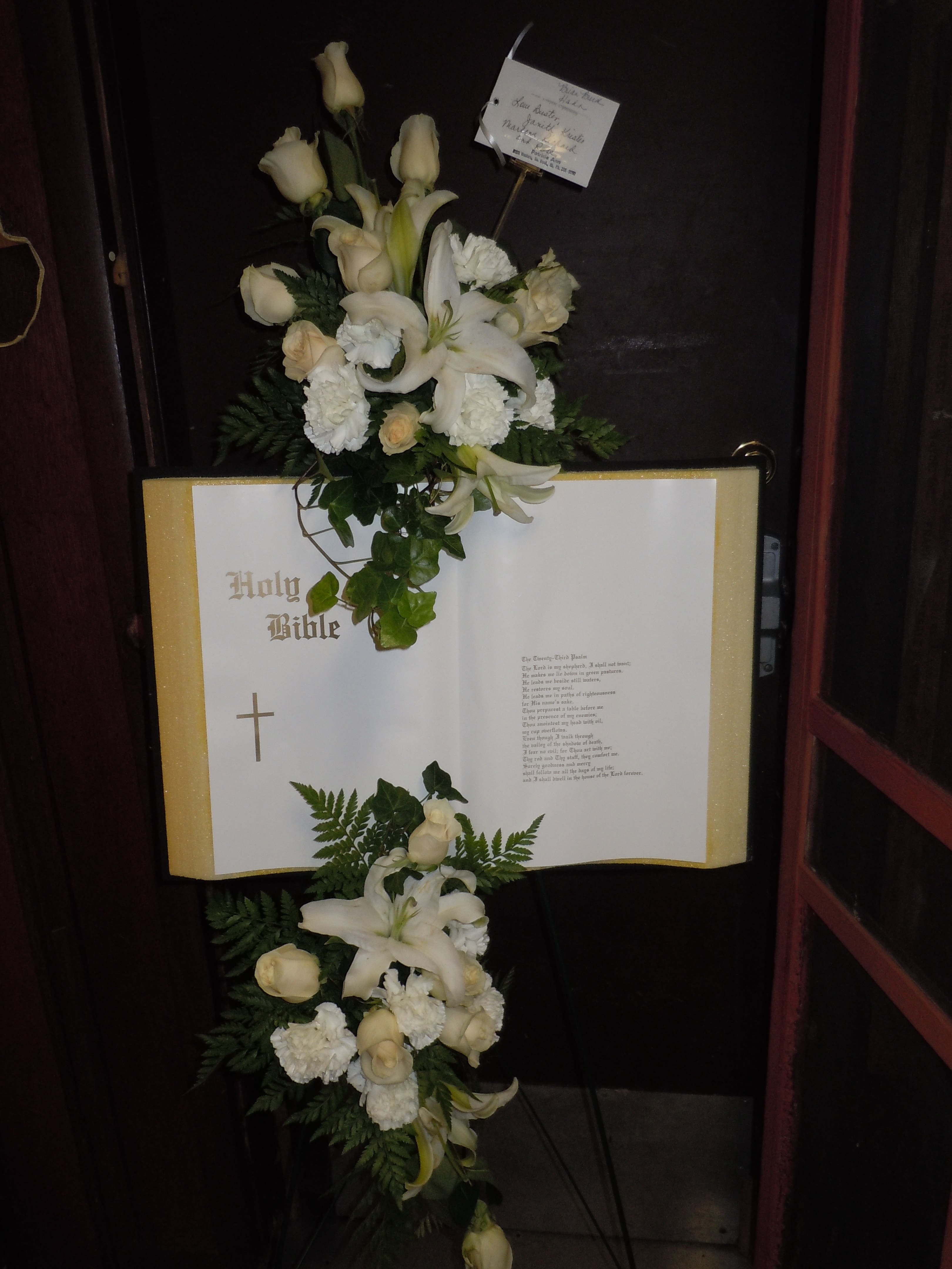 SP33 Standing Spray - A mixture of white carnations, white roses, lilies  and greens with the stryofoam bible.  Banner - $15.00 + extra   Substitutions may apply due to flower availability