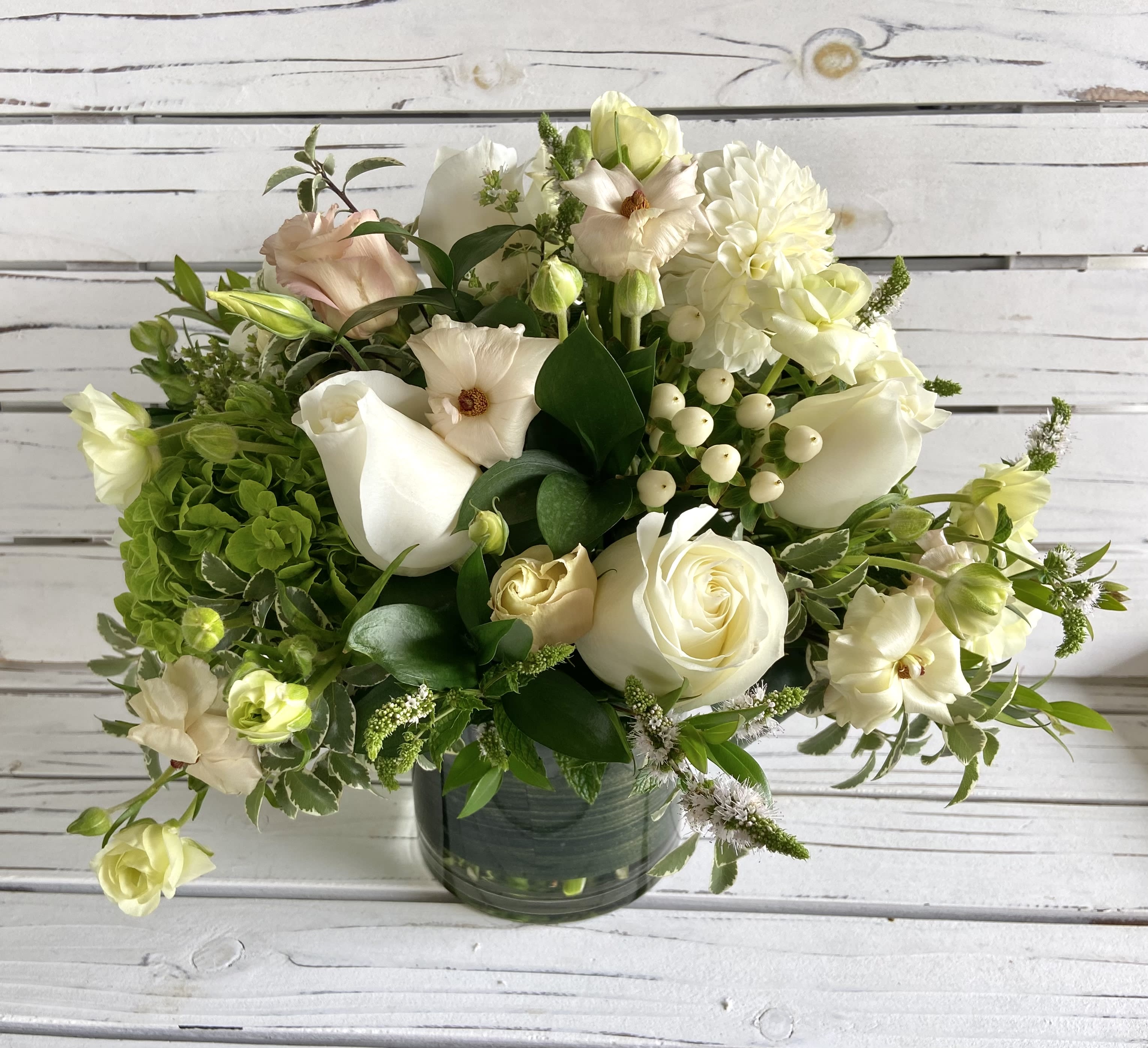 A Touch of Blush - Whites and greens with a touch of blush In a 5x5 cylinder. Approximately 14 inches around and tall. Exact flowers and vase may vary. 