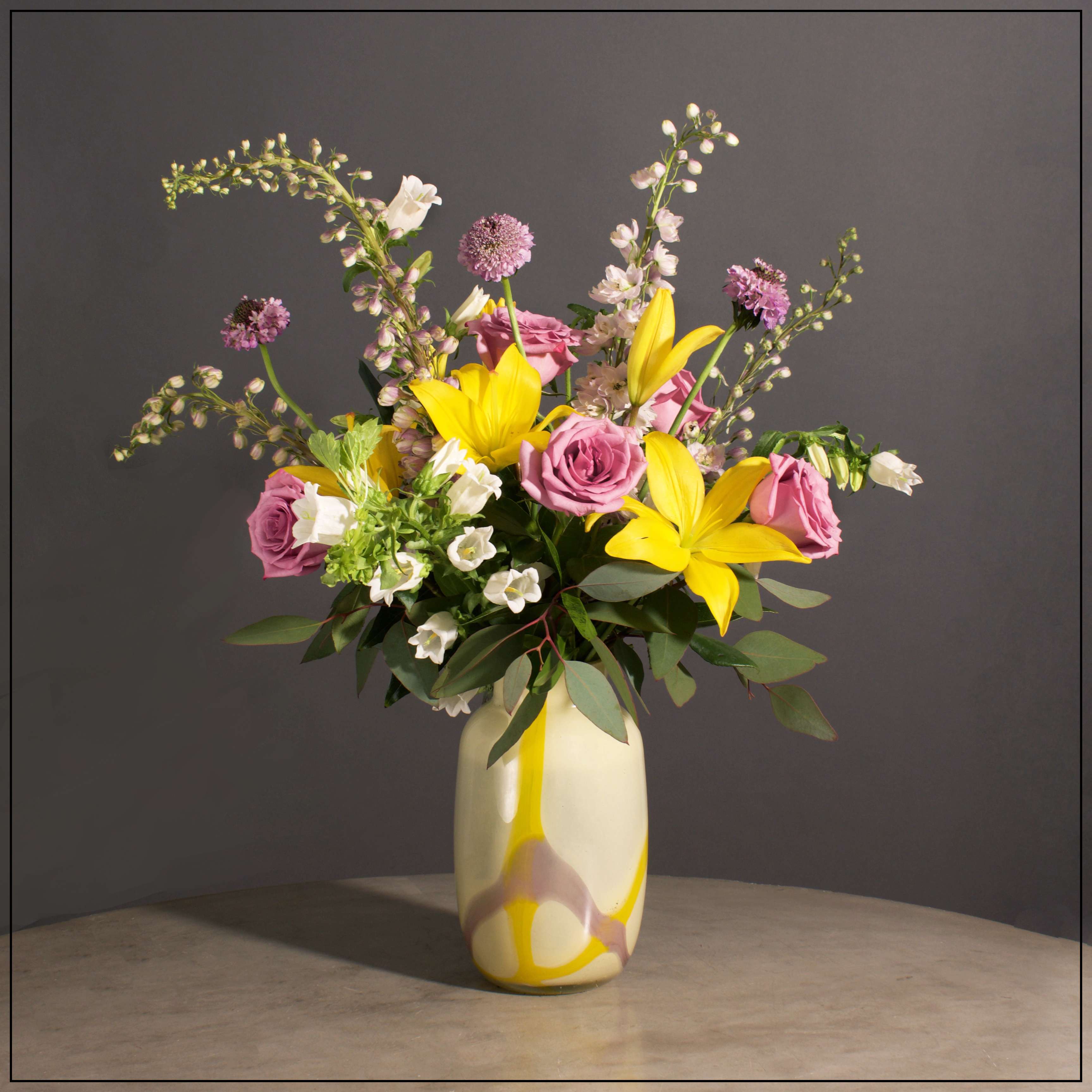 Portofino - Delphinium, Lillies, Bells of Ireland, Campanula, Roses and Scabiosa with Eucalyptus in a Beautiful Art Glass Vase.  Approximately 17" H x 23" W