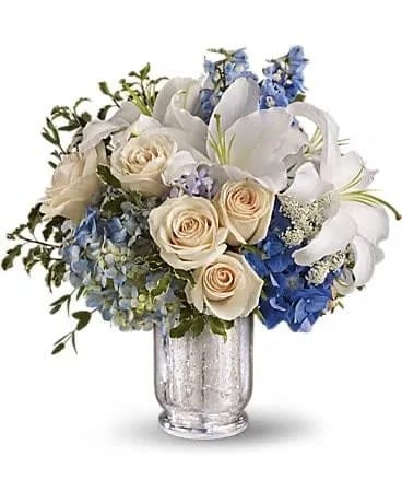Seaside Centerpiece - Spend a peaceful afternoon at the seaside with this breathtaking centerpiece. Lush blue hydrangea and delphinium evoke sea and sky, while creamy roses, white lilies and delicate Queen Anne's lace whisper of sand and sea foam. Presented in an antiqued Mercury Glass Vase, it's an elegant choice for weddings and showers. Two shades of blue hydrangea are mixed with crÃ¨me roses, light blue delphinium, fragrant white oriental lilies, delicate Queen Anne's lace, parvifolia eucalyptus and green pitta negra in a beautiful Mercury Glass Antique Hurricane. 