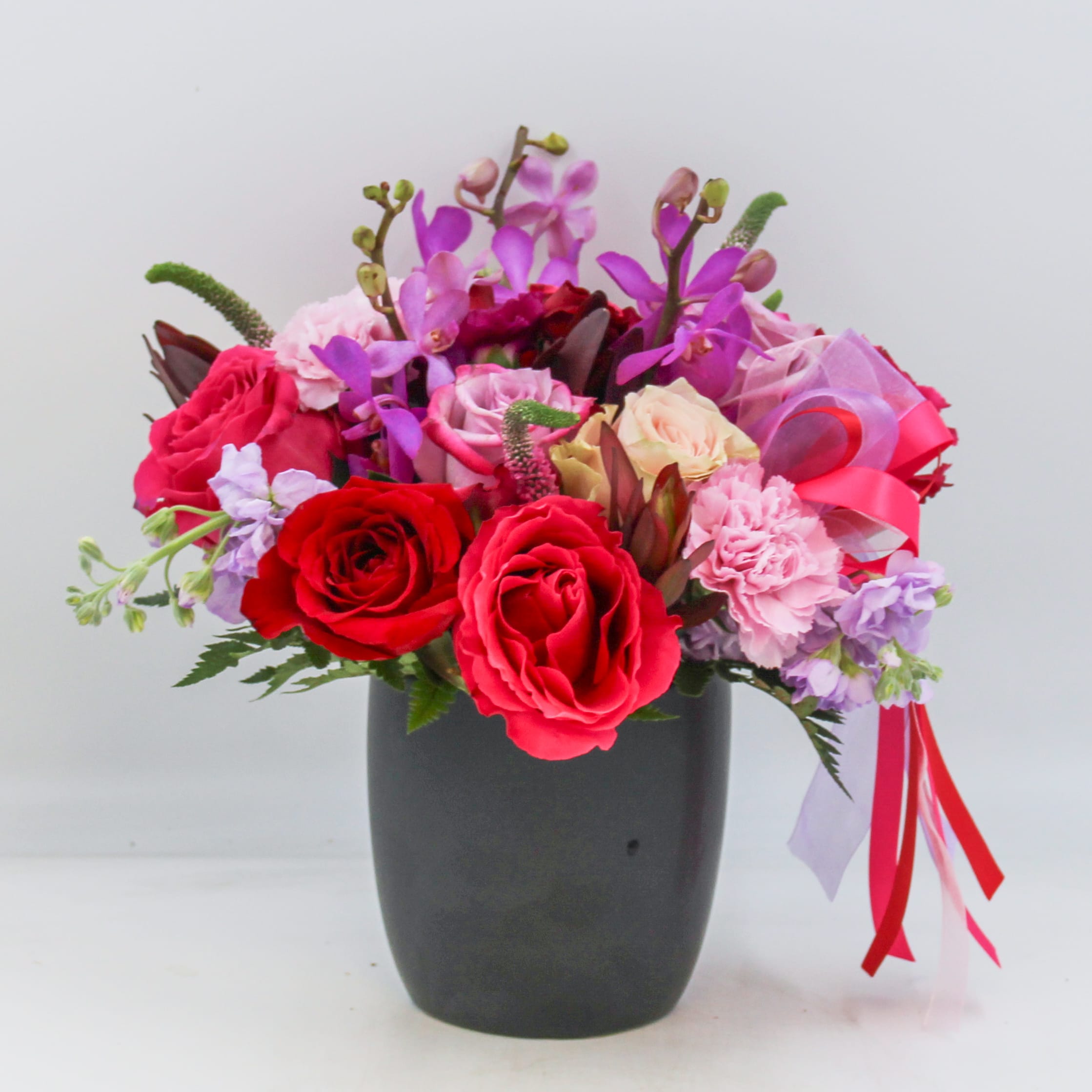 London  - London is a limited edition design for our Valentine’s en vogue collection and features Lecadendron and Mokara Orchid tropicals, Lavender Stock, Red Roses, Blush Roses, Lavender Roses, Hot Pink Roses, Pink Veronica, Red Spray Roses and Pink and Purple Carnations arranged in a black keepsake ceramic pot adorned with mixed colored satin and chiffon ribbons. 