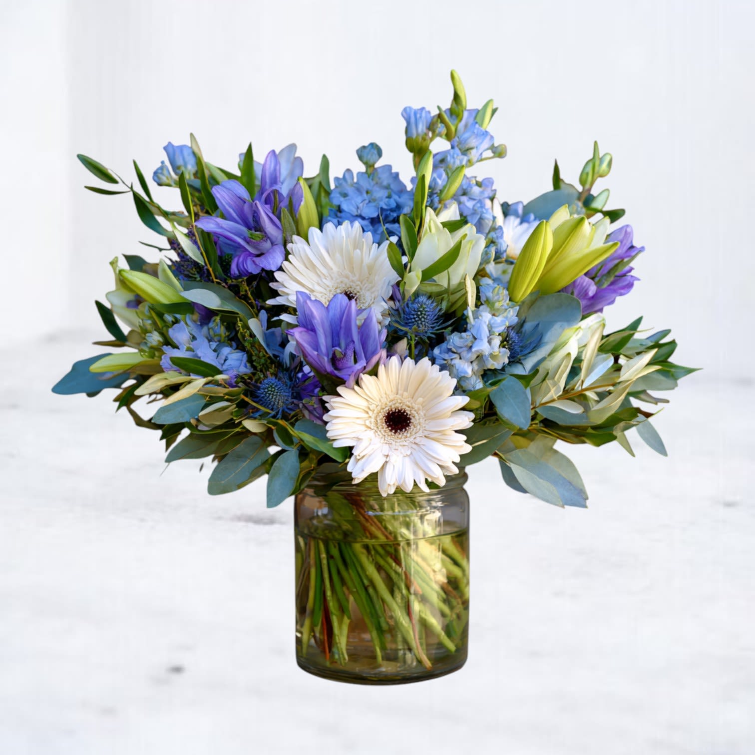 Liora - Liora is a light-filled arrangement designed to convey peace, reassurance, and quiet beauty. Soft blue florals, creamy white blooms, and fresh eucalyptus are gathered in a relaxed, garden-inspired style that feels calm and comforting without formality.  Thoughtfully balanced and effortlessly elegant, Liora is a meaningful choice for sympathy gestures, moments of support, or simply sending something serene and uplifting.