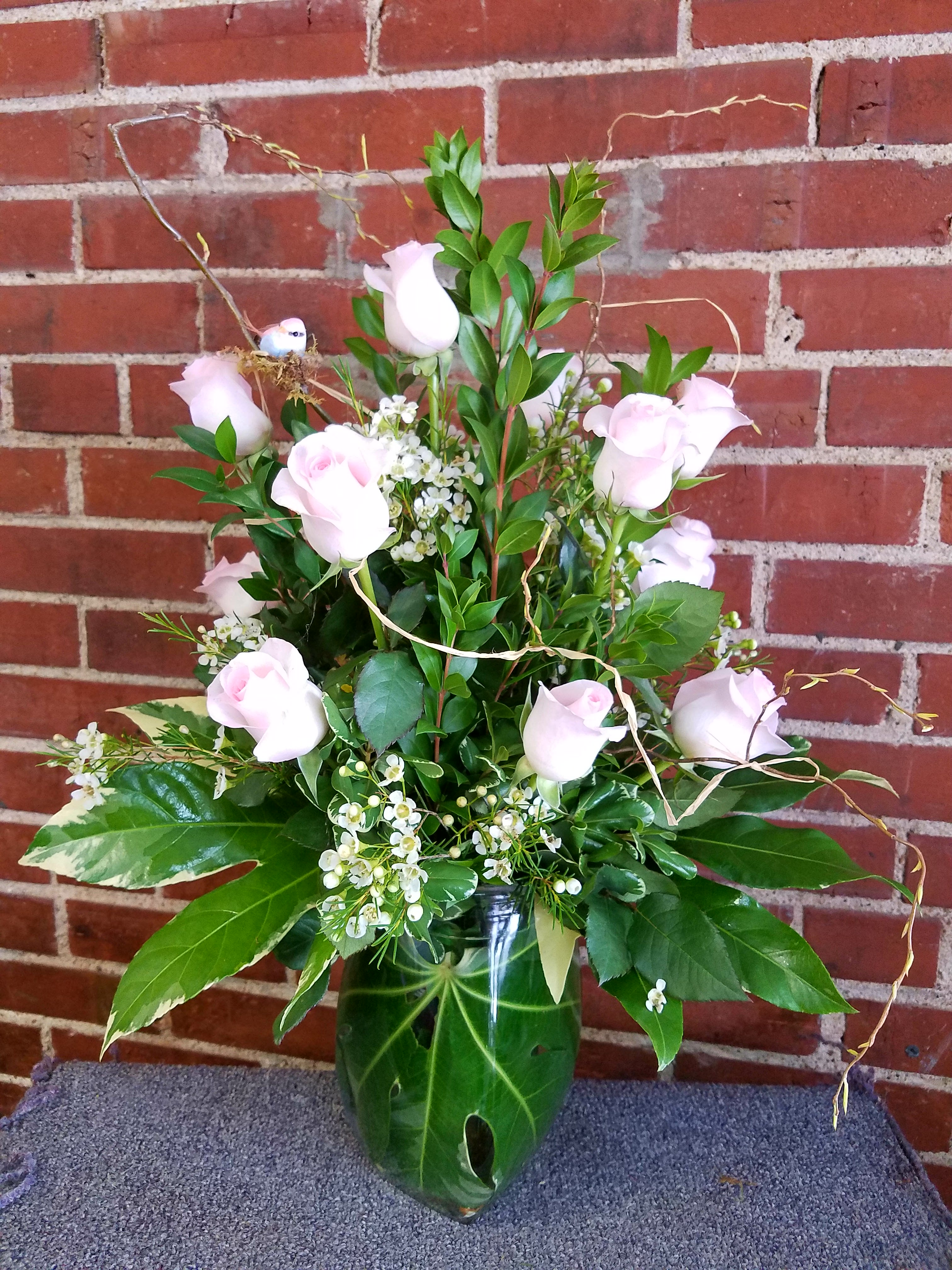 Eckert Florist's Pink Rose Bouquet - The Pink Rose Bouquet is a graceful expression of blushing beauty. One dozen of our finest pink roses are perfectly accented with lush greens, floral filler, and arranged in a clear glass vase to create a sentiment of cheerful wishes that will bring comfort and warmth. *Shade of Pink Rose Will Vary