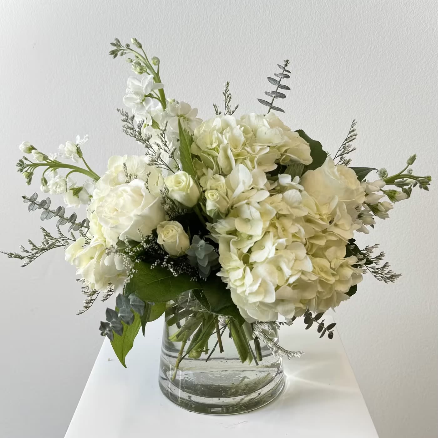 Simply Pure - Discover the serene elegance of Simply Pure, an exquisite all-white flower arrangement that radiates timeless sophistication and tranquil beauty. This luxurious bouquet showcases lush white hydrangeas as its romantic centerpiece, their full, cloud-like blooms creating a soft, dreamy foundation. Velvety white roses — the classic emblem of purity, love, and new beginnings — add classic charm and delicate fragrance, while dainty white spray roses bring subtle texture, gentle volume, and effortless grace throughout the design. Tall, graceful white stock introduces elegant vertical lines and a sweet, honey-spiced scent that lightly perfumes the air. Fresh, silvery-green eucalyptus leaves provide the perfect natural contrast, their aromatic, rounded foliage framing the pristine white flowers with depth, movement, and a touch of wild sophistication. Ideal for weddings, bridal bouquets, sympathy arrangements, anniversaries, new baby congratulations, or any moment calling for understated luxury, Simply Pure embodies minimalist chic and peaceful serenity. Handcrafted by expert florists using the freshest premium blooms, this all-white floral masterpiece arrives ready to inspire calm, joyful elegance. Perfect for those seeking clean, modern floral designs, pure white hydrangea bouquets, elegant white rose arrangements, or sophisticated monochromatic flowers that whisper refined simplicity and heartfelt emotion.