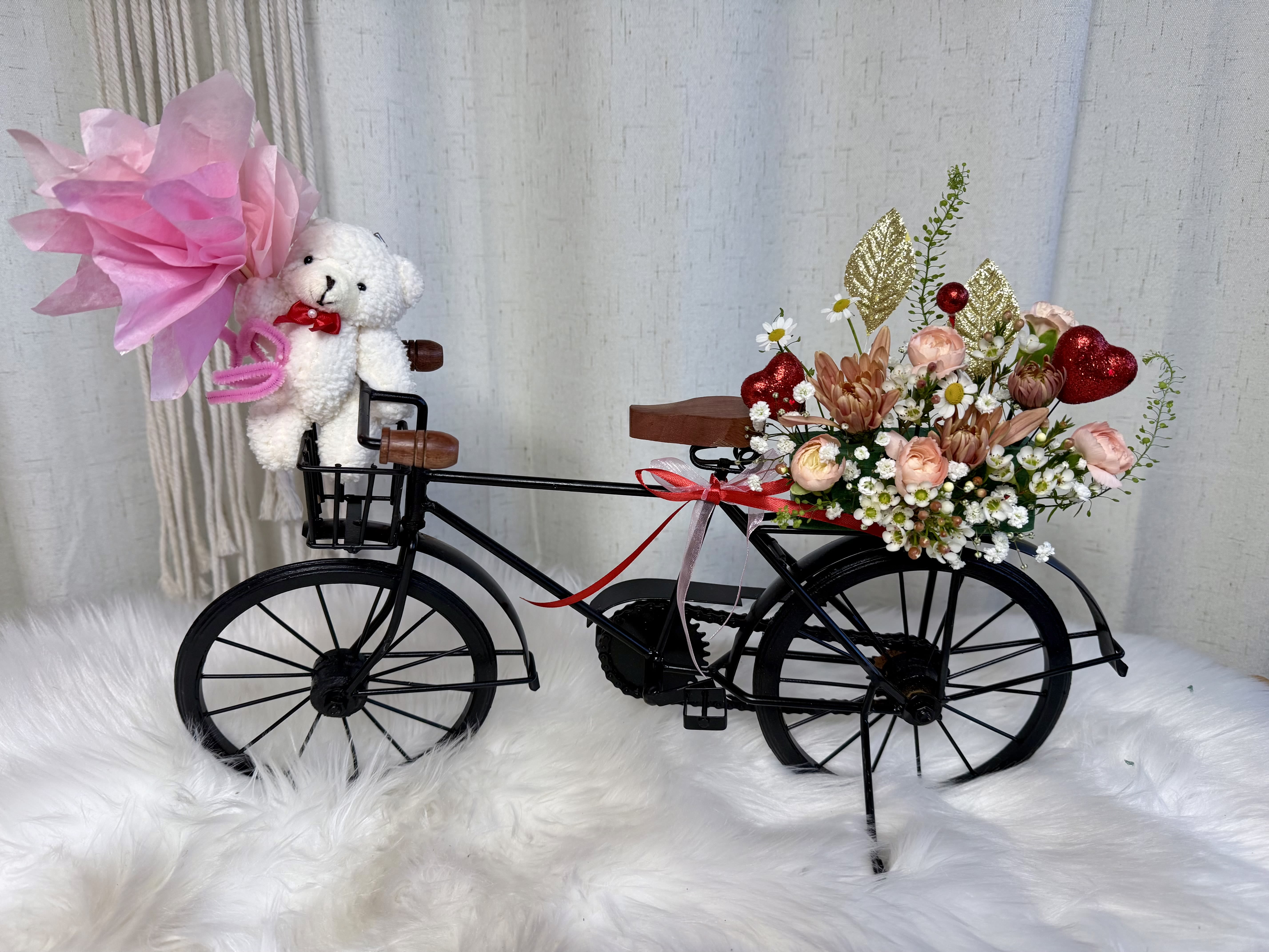 Take me on your bicycle. - A gift full of love and sweetness . Our decorative bicycle with teddy bear and floral arrangement is perfect for surprising someone special and bringing a smile to their face. Each piece is carefully designed with love, combining delicate flowers and an adorable teddy bear that adds charm and elegance.   Available in two bicycle colors: Black and Gold   Please note: When placing your order, kindly let us know in the “special instructions” which bicycle color you prefer.  Aproximadamente 1 pie de alto y 1,8 pulgadas de ancho. 