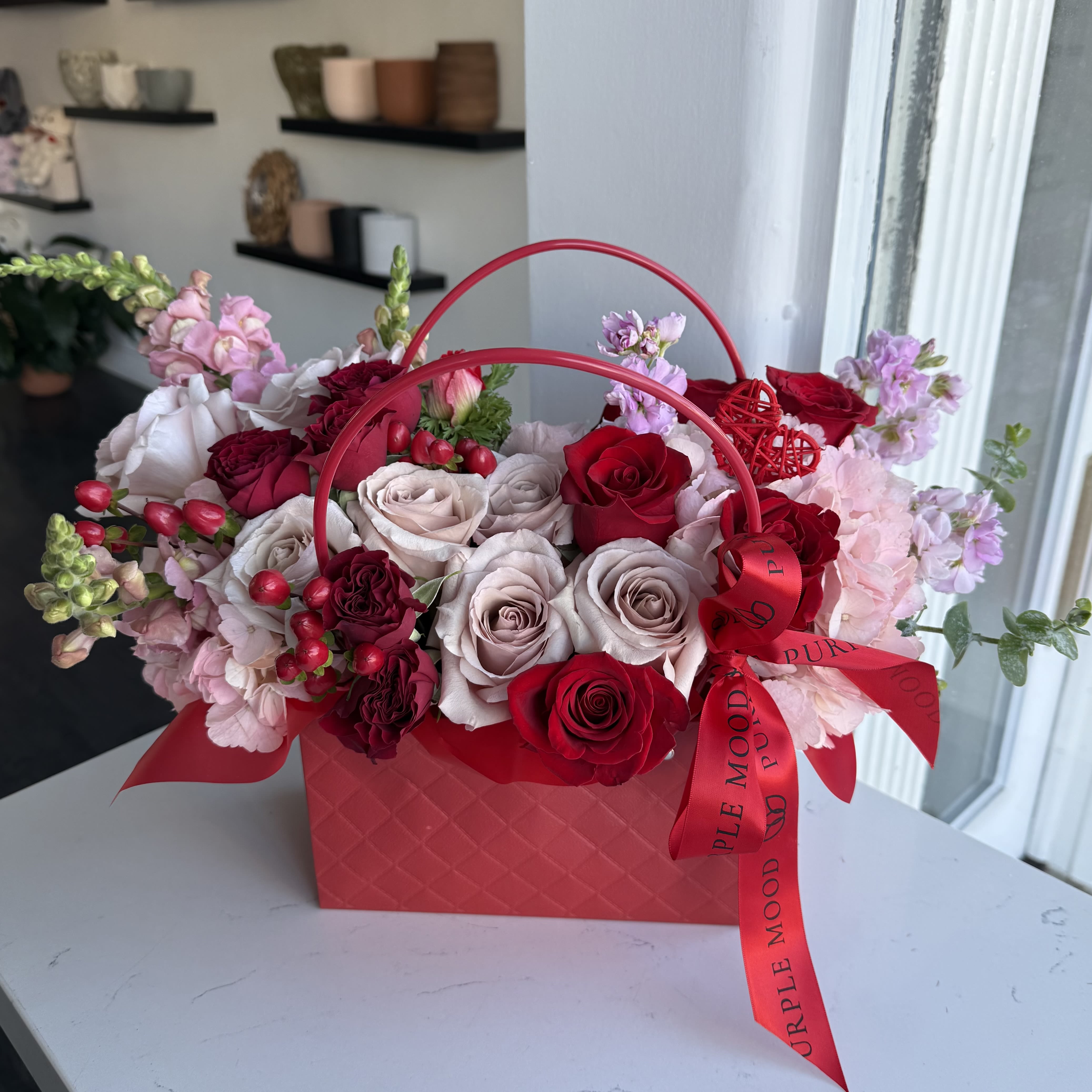 Red mini Bag - Description A chic and romantic floral handbag arrangement featuring premium red and blush roses, soft hydrangea, seasonal blooms, and vibrant berry accents, designed in a signature red mini bag with Purple Mood’s iconic ribbon. This stunning Valentine’s piece blends luxury, romance, and playful elegance — the perfect gift to say “Be My Valentine.”  Perfect for girlfriends, wives, and anyone who loves modern, fashion-inspired florals.   Handcrafted by Purple Mood Flowers  Limited Valentine’s Edition