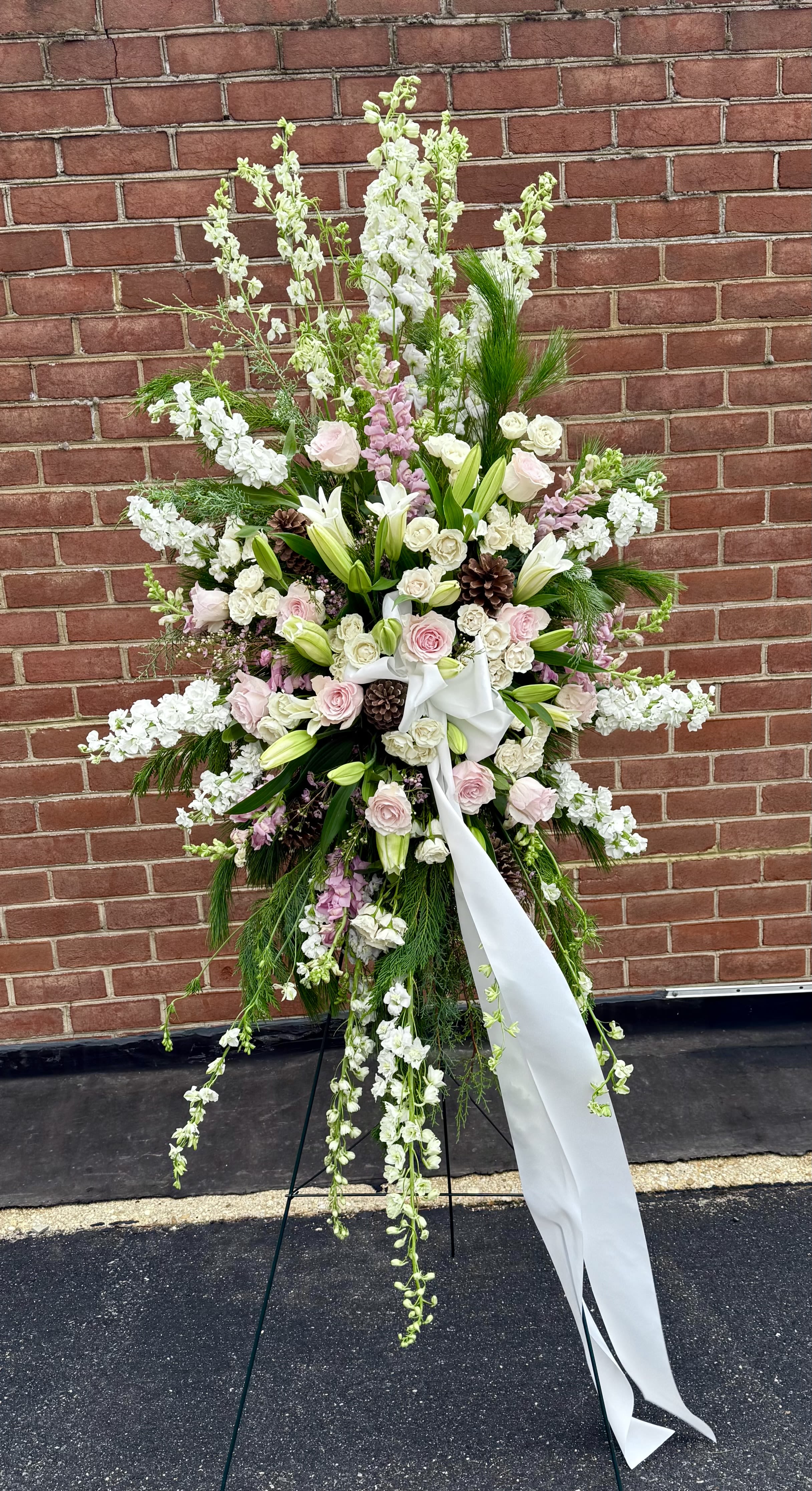 Peaceful Remembrance - An elegant sympathy easel arrangement featuring soft white lilies, roses, and seasonal blooms accented with gentle blush tones and lush greenery. Designed with graceful height and flowing textures, this standing spray conveys peace, remembrance, and heartfelt condolences, offering a beautiful tribute to honor a loved one’s life.  (The greenery will vary this is s seasonal arrangement.)