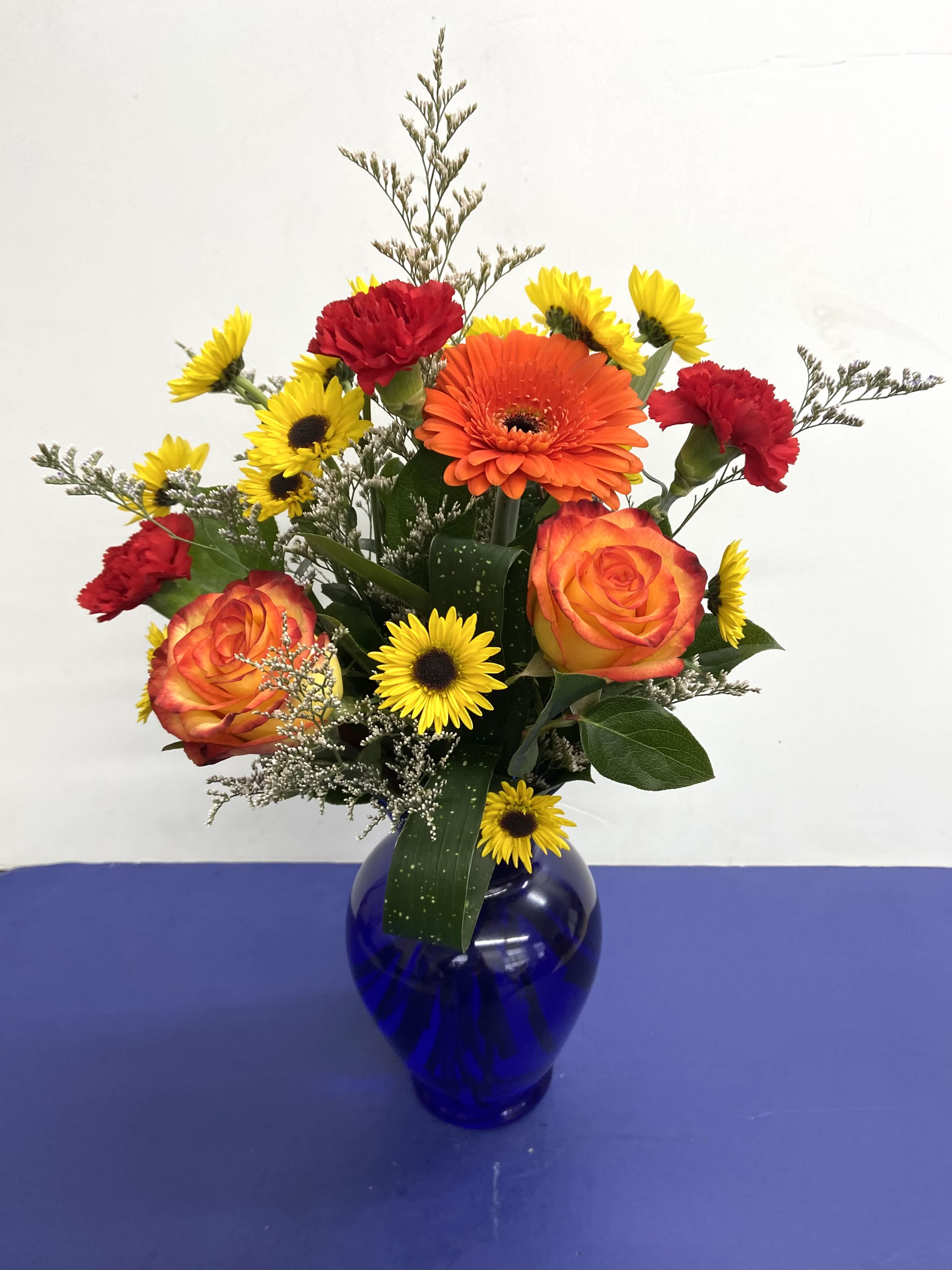 Fly Away Birthday - Brilliant orange roses, red carnations and yellow daisy chrysanthemums, purple statice delivered in a beautiful cobalt blue vase.    