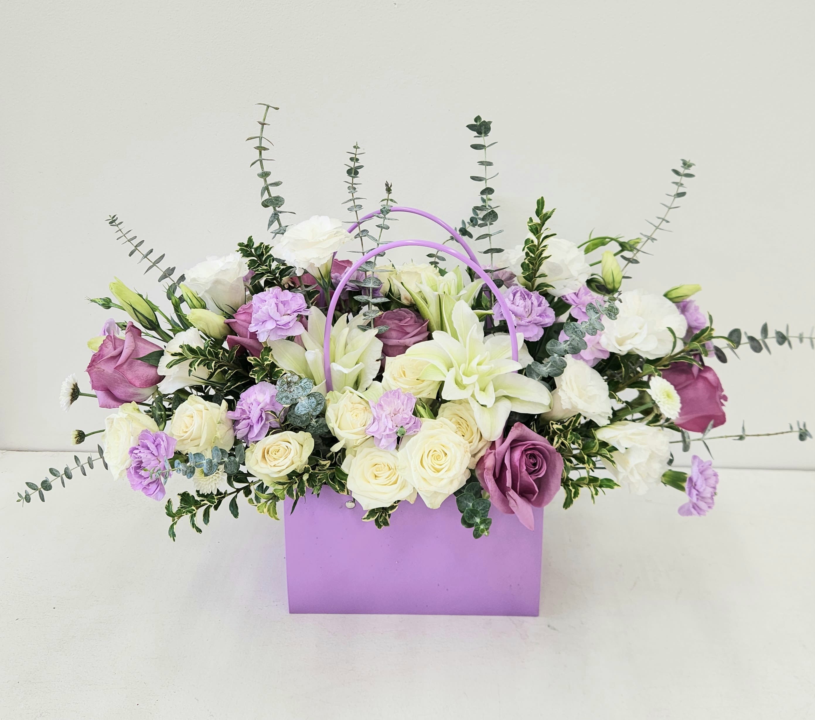 Bixby Charms - An elegant and unique bouquet of fresh roses and lilies, beautifully arranged in a lavender gift bag with a handle for easy carrying. A charming blend of style and convenience, perfect for gifting or brightening any day.  AS PICTURED: PREMIUM 18"W by 18"H
