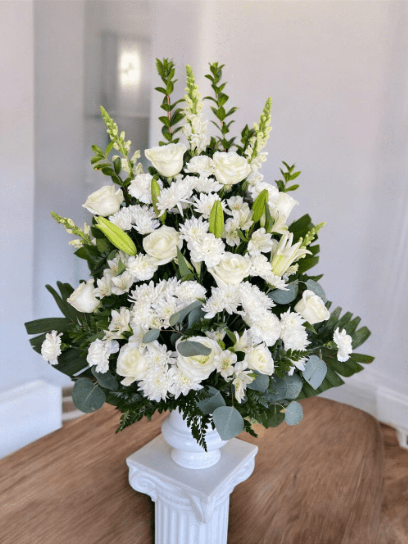 Classic White Arrangement  - An elegant and serene floral arrangement designed with a harmonious blend of white roses, chrysanthemums, lilies, and delicate greenery. This classic white arrangement conveys respect, comfort, and hope, making it a thoughtful choice for expressing sympathy, honoring loved ones, or offering a message of love and tranquility during difficult moments. Presented in a classic white base that enhances its timeless beauty and dignity.   Un arreglo floral de gran elegancia y serenidad, diseñado con una armoniosa combinación de rosas blancas, crisantemos, lirios y delicado follaje verde. Este arreglo clasico blanco transmite respeto, consuelo y esperanza, siendo una elección ideal para expresar condolencias, acompañar homenajes o brindar un mensaje de amor y tranquilidad en momentos difíciles. Presentado en una base clásica blanca que realza su belleza y solemnidad.  