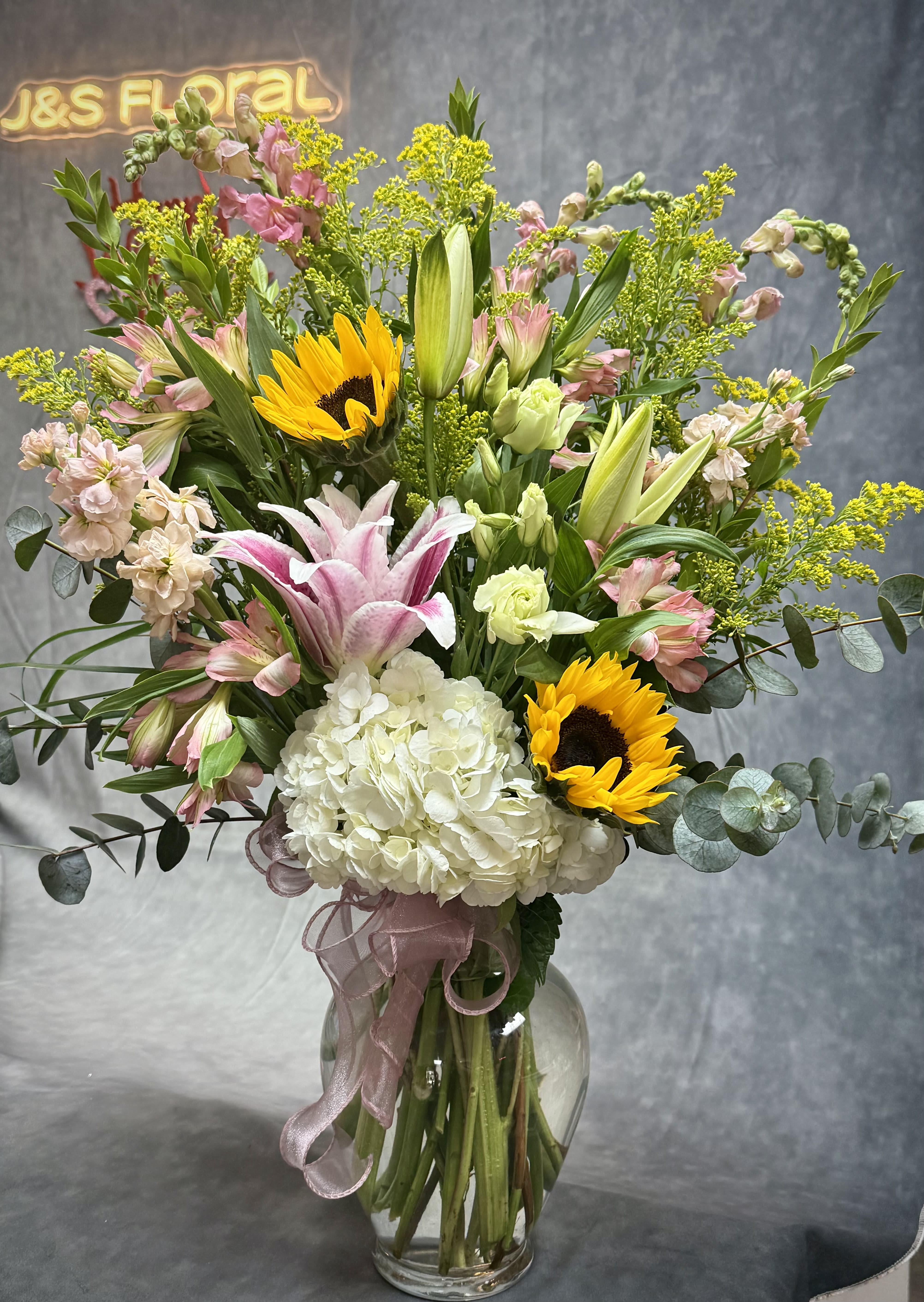 Golden Garden Symphony  - A radiant burst of elegance featuring cheerful sunflowers, lush hydrangeas, graceful lilies, striking calla lilies, Bells of Ireland, and seasonal blooms—all artfully arranged in a designer vase. This vibrant creation brings the beauty of a blooming garden right into your space. 