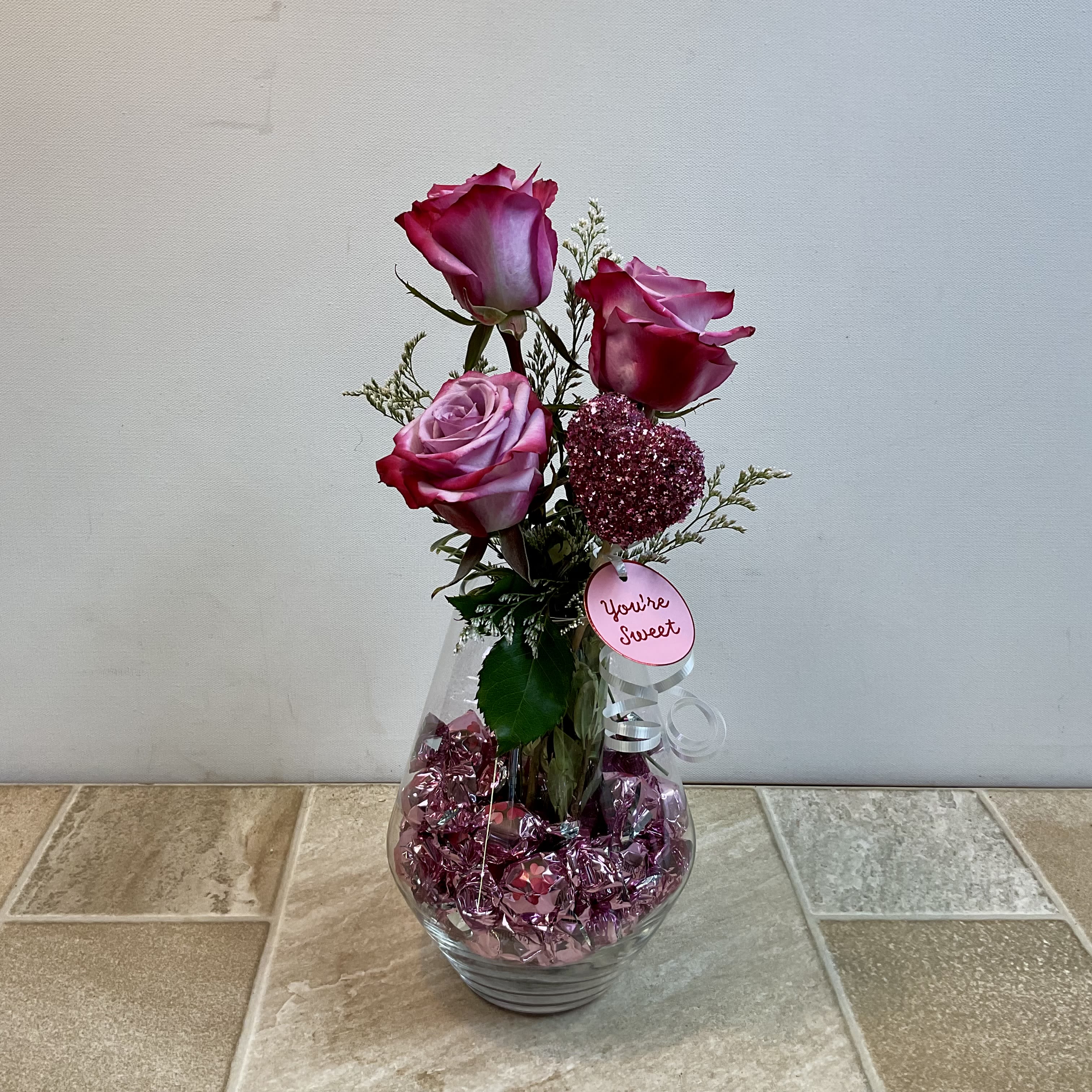 You’re Sweet - This fresh flower and candy bouquet is a charming blend of romance and sweetness. Three elegant lavender roses—each with softly blended blush and raspberry tones—rise gracefully from a clear, rounded glass vase. Delicate wisps of airy greenery add lightness and texture, while a sparkling pink heart accent brings a playful, love-filled touch.  Nestled inside the vase are shimmering pink foil-wrapped chocolates, creating a candy-filled base that enhances the bouquet’s festive feel. A petite tag reading “You’re Sweet” finishes the design, making the message unmistakably heartfelt. Overall, this arrangement feels flirty, thoughtful, and perfect for Valentine’s Day, anniversaries, or any occasion meant to say “I care.”   Primary flowers: rose  Flower delivery La Crosse, Onalaska, Holmen, West Salem, La Crescent. Flower shop. Plant store. Best florist La Crosse Wisconsin area.  Seasonal and holiday floral creations. Funeral arrangements. New baby bouquets. Birthday celebration flowers. Get well arrangements. Just because gifts.