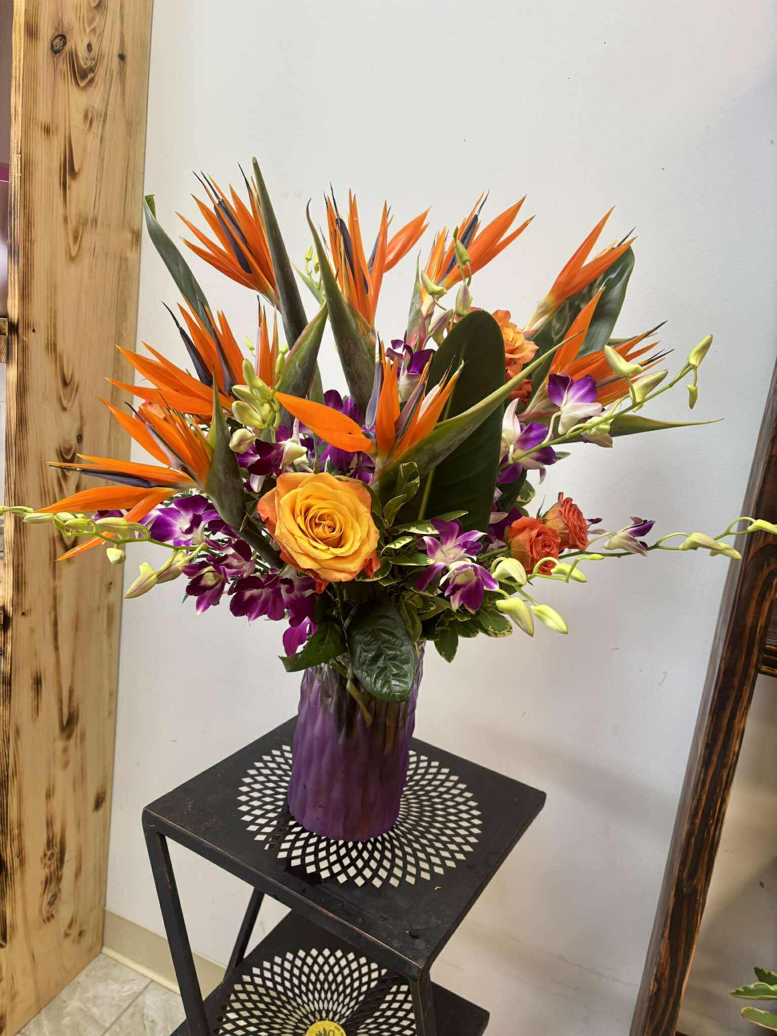 Tropical Sunset Arrangement - Bring a burst of paradise into any space with our stunning Tropical Sunset Arrangement. Designed in a rich purple vase, this vibrant display features the dramatic beauty of Birds of Paradise, paired with bright orange roses and exotic purple dendrobium orchids.  Perfect for birthdays, celebrations, or simply adding a touch of the tropics to your home or office, this arrangement is sure to impress.