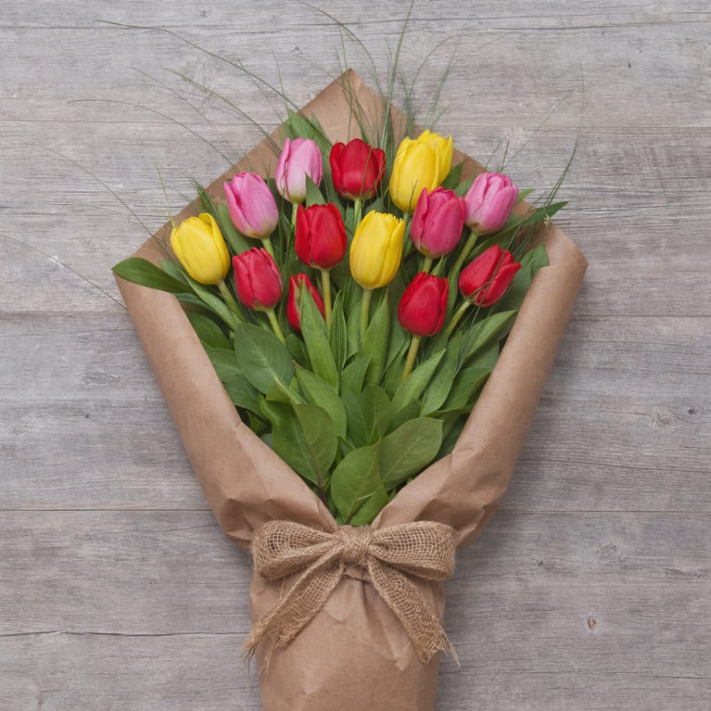 Spring Tulip Medley - A vibrant wrapped bouquet of fresh tulips in cheerful shades of pink, red, and yellow, complemented by seasonal greenery and wispy grass accents. Finished in rustic kraft paper with a burlap bow, this bouquet captures the joy and simplicity of spring—ideal for birthdays, thank-yous, or just because.